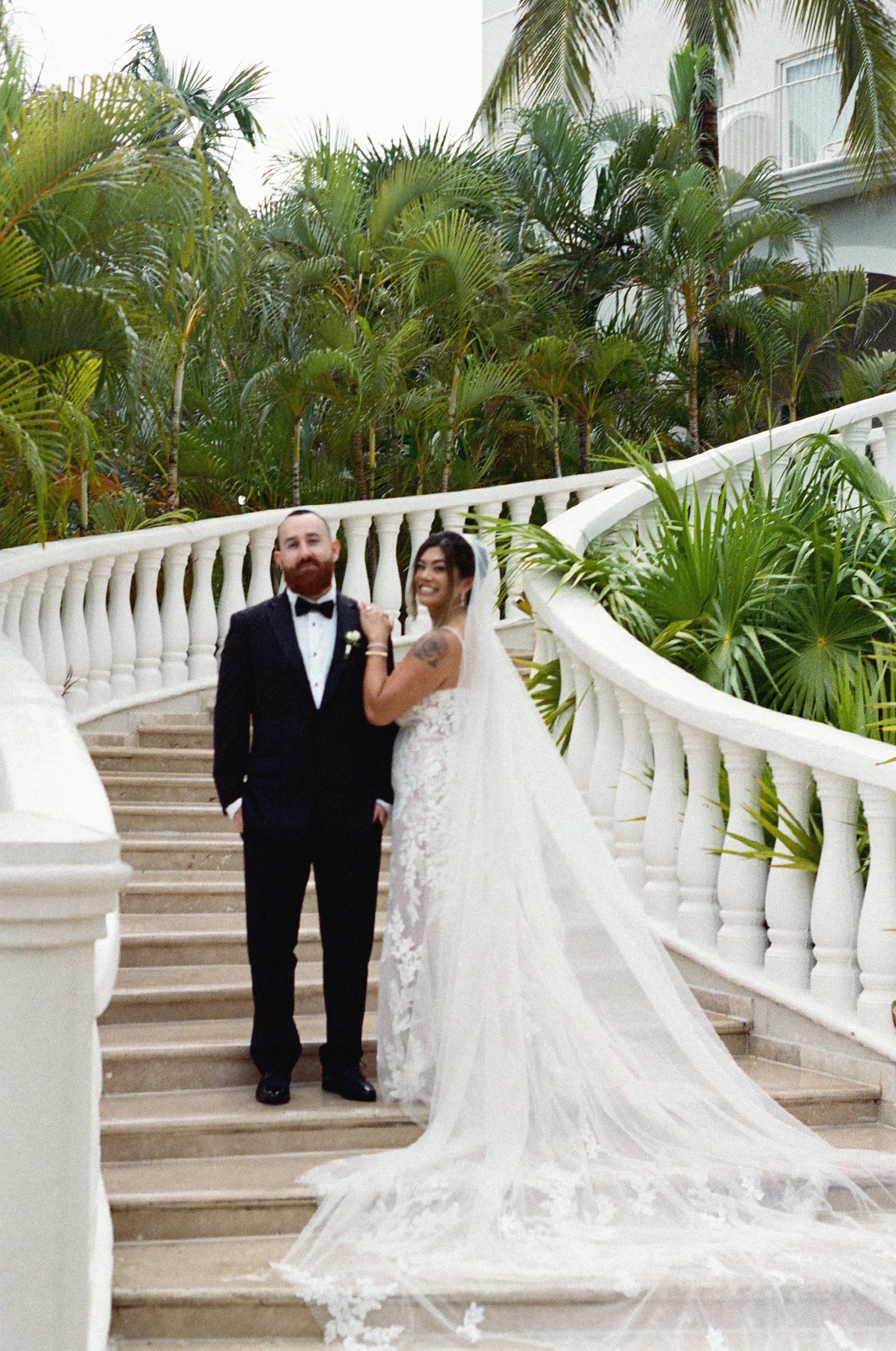 A bride and groom standing on a staircase with white balustrades and lush green plants in the background.