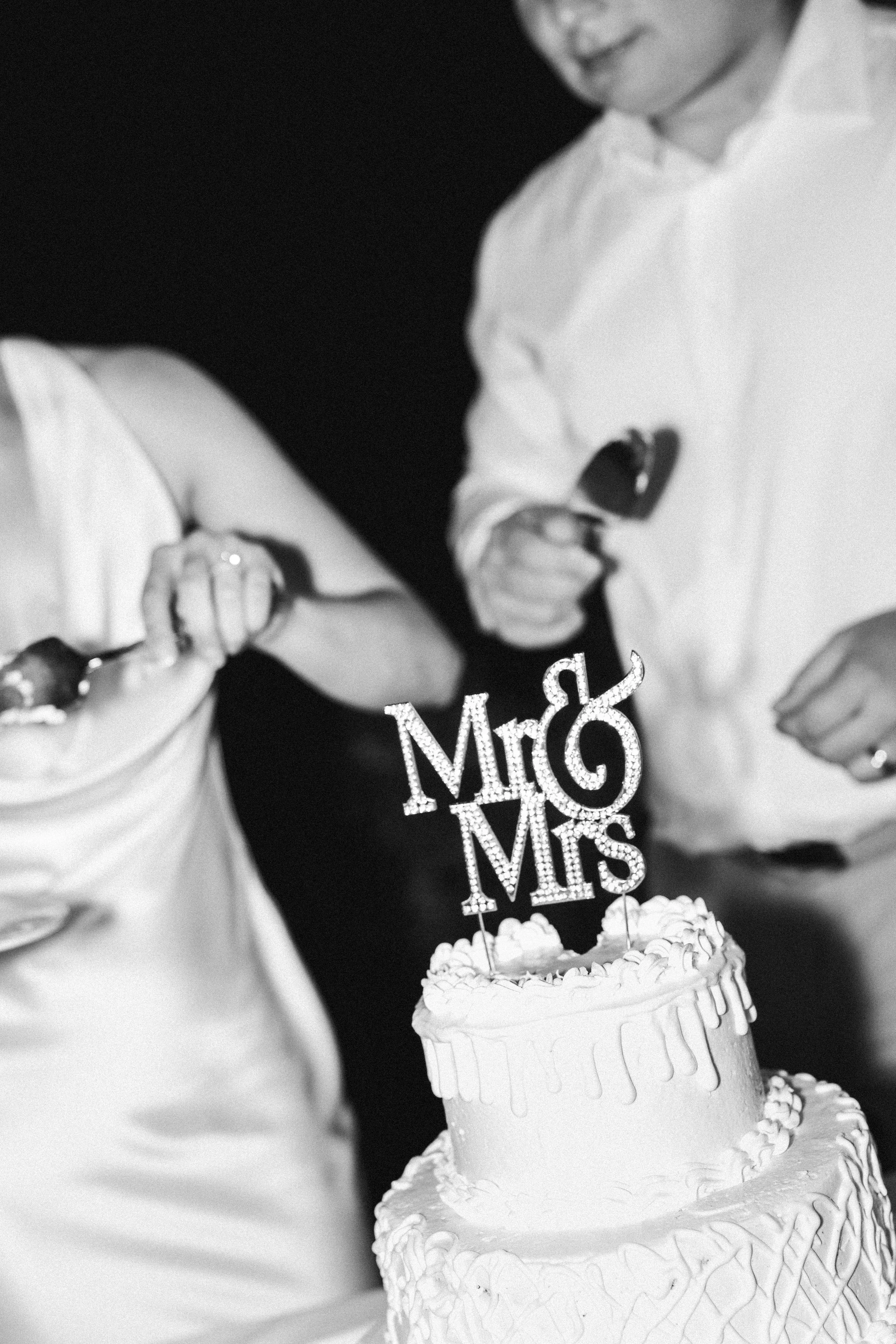 A wedding celebration scene with a two-tiered decorated cake, and bride and groom cutting their cake. 'Mr & Mrs' topper. People are gathered around, and one person appears to be cutting the cake.