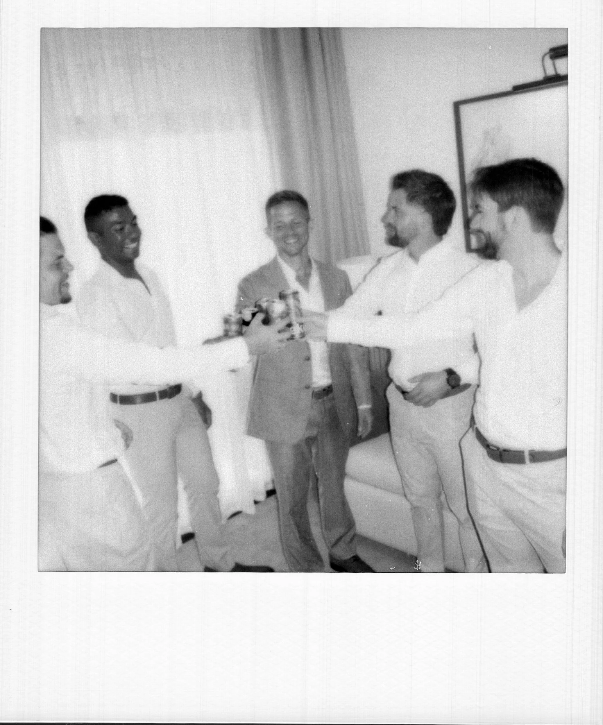 A group of five men standing in a semi-circle indoors, engaging in a toast with small glasses, smiling, with a framed picture on the wall behind them.