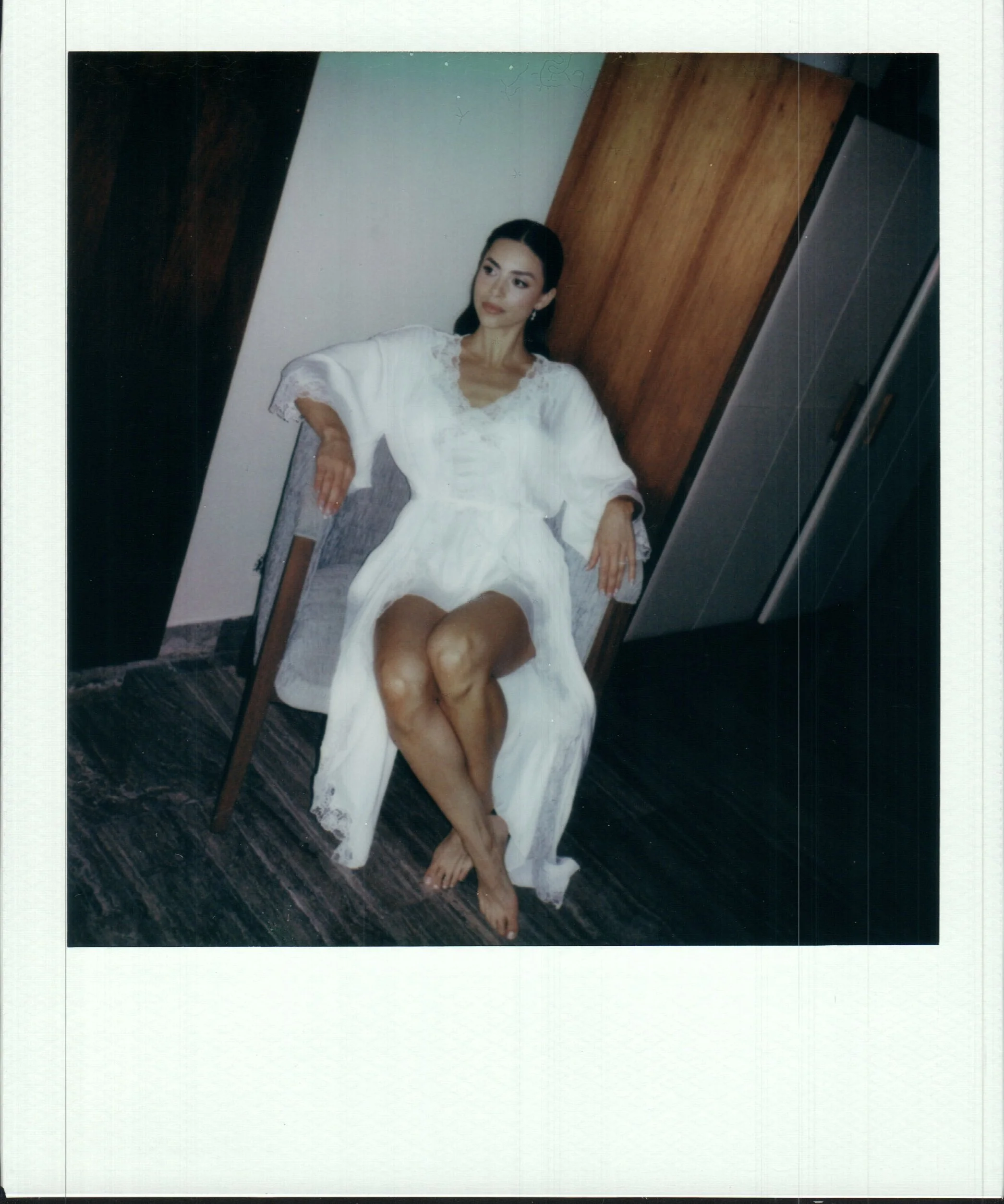 A woman in a white dress sitting on a chair with her legs crossed and her arm resting on the back of the chair, against a background of dark wood paneling and a white wall.
