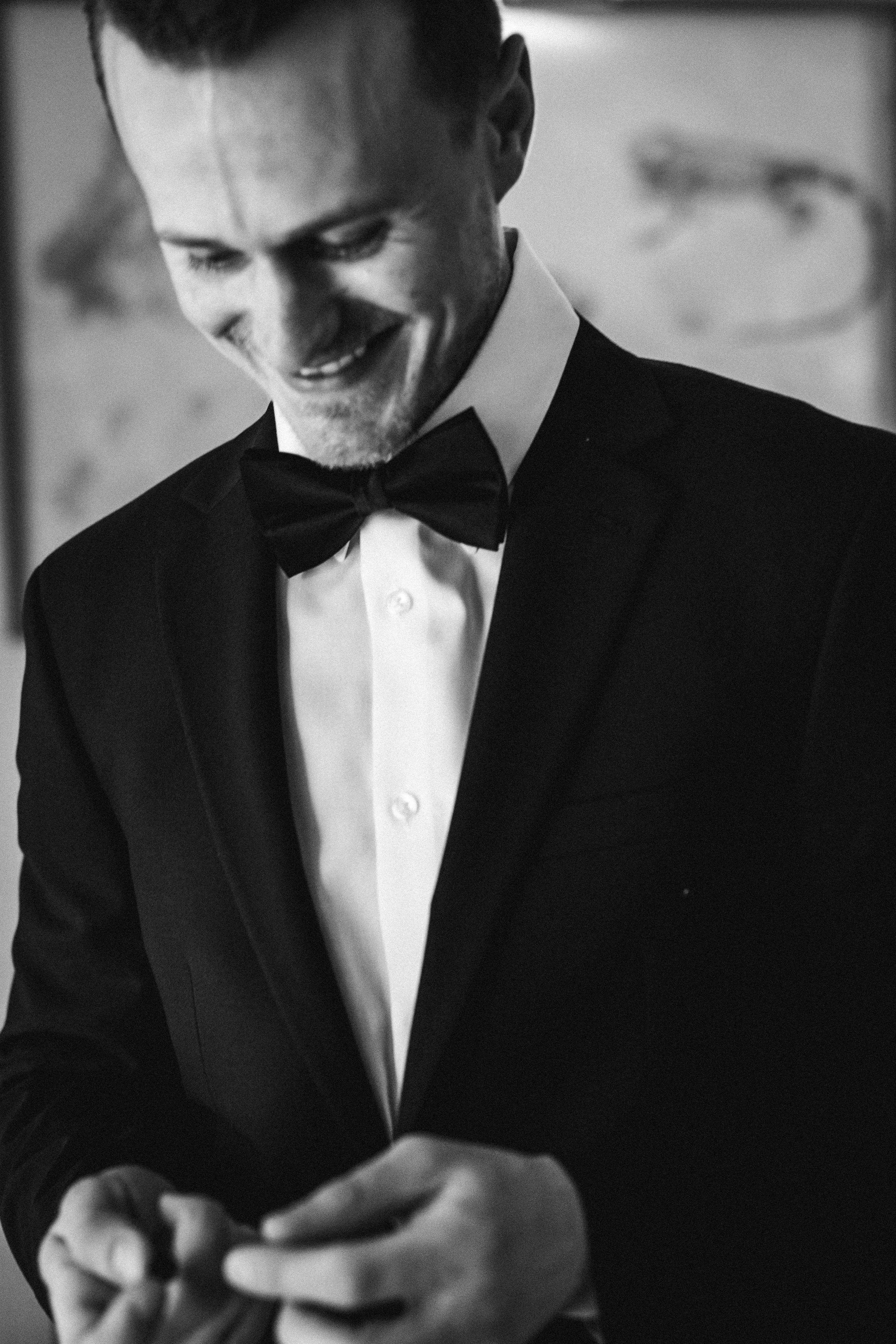 A groom in a tuxedo with a bow tie, smiling and looking down, appears to be adjusting his cufflinks.