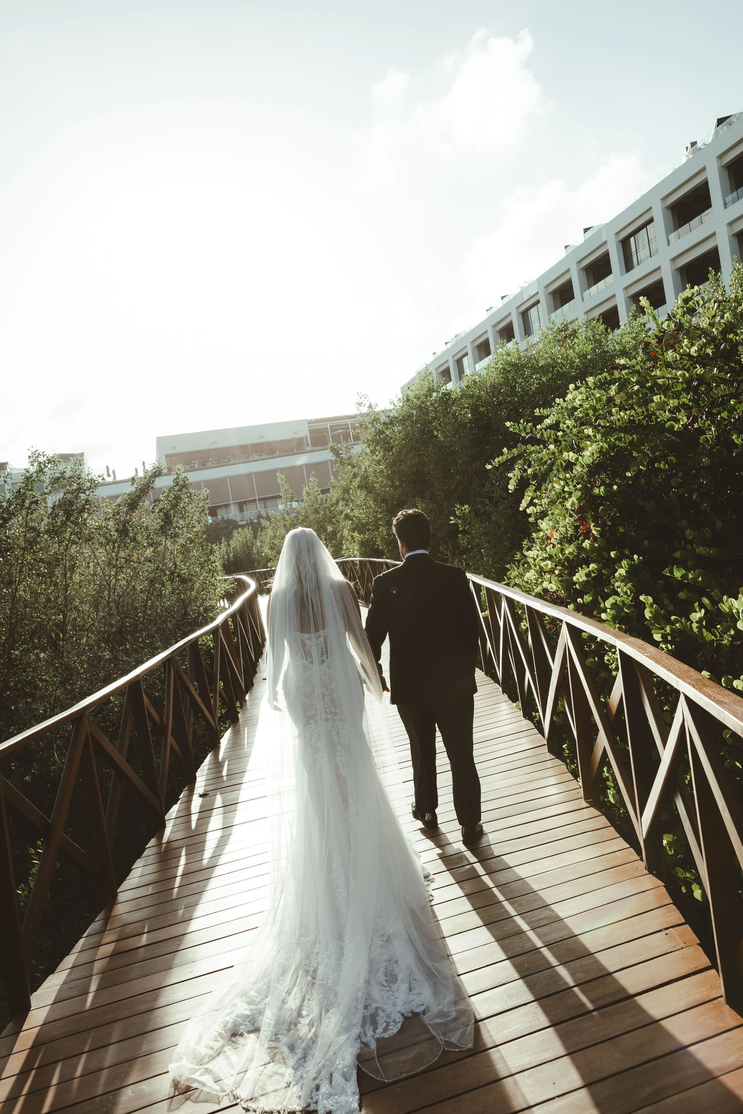 Bride and groom walking hand in hand on a wooden bridge outdoors, surrounded by greenery, with modern buildings in the background, under a partly cloudy sky in Atelier Playa Mujeres Wedding.