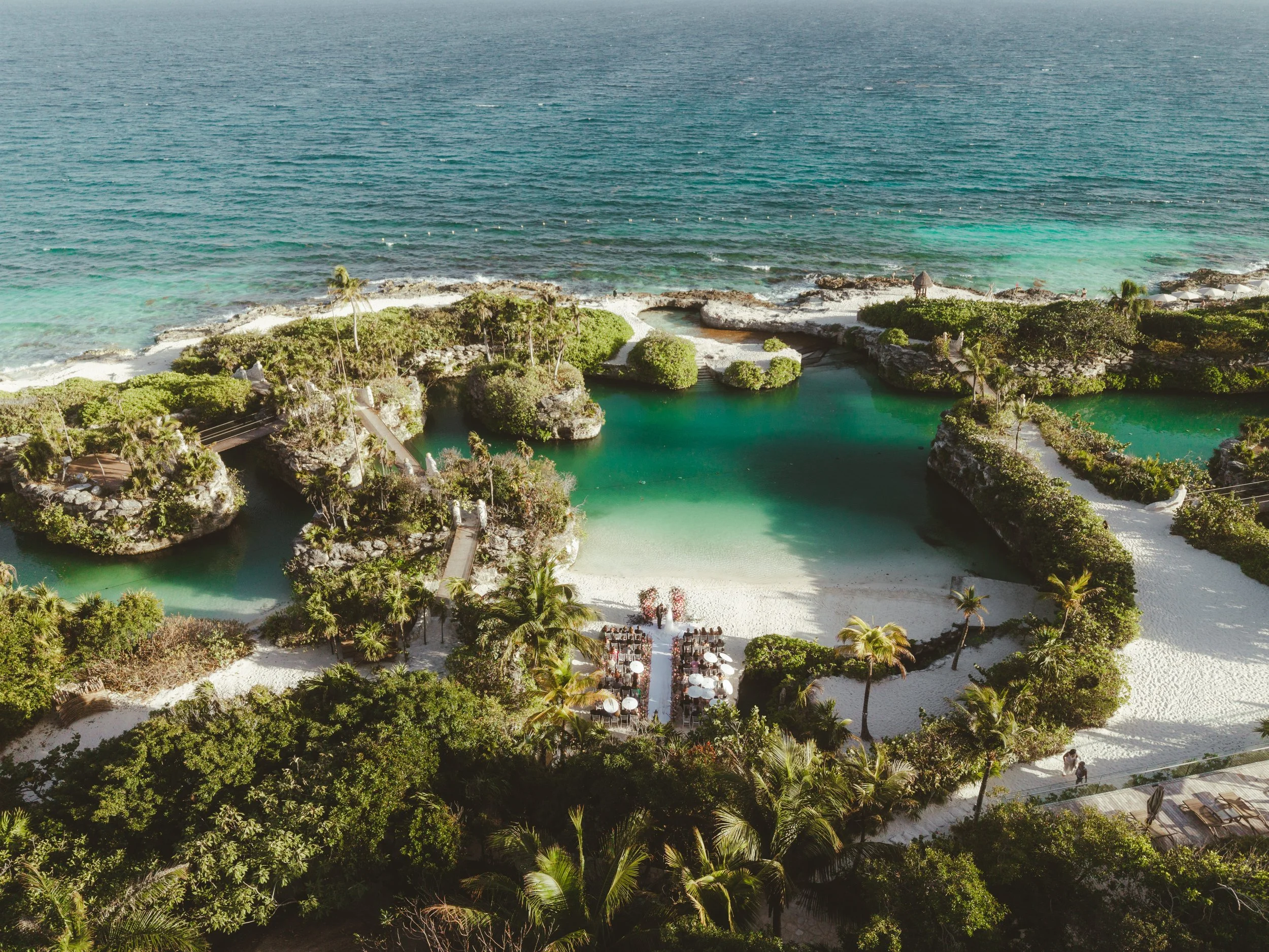 Aerial view of a tropical seaside resort wedding venue set up, with green pools, lush trees, sandy paths, and ocean waves in the background. Xcaret Mexico Wedding.
