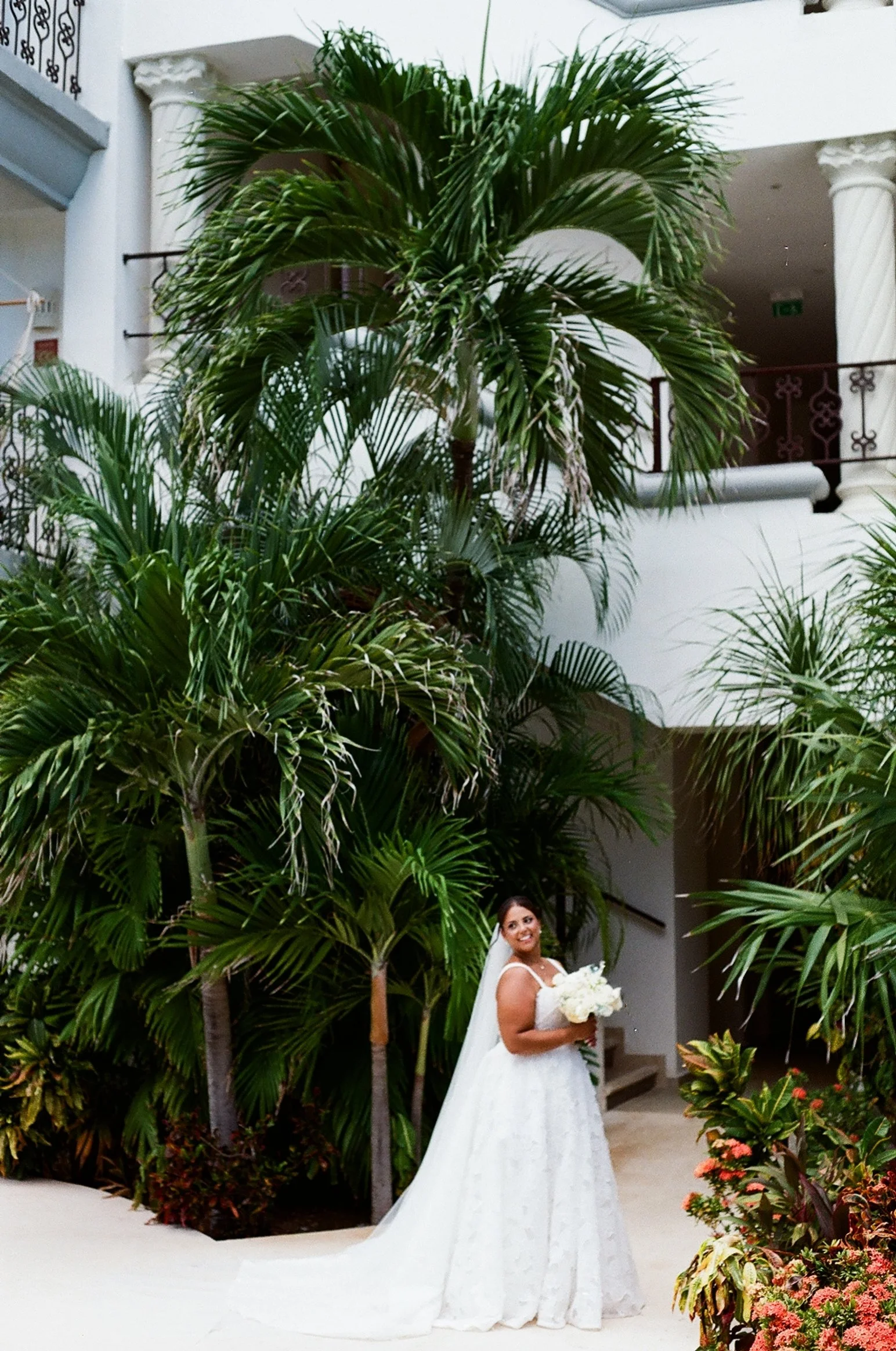 A bride in a white wedding dress holding a bouquet of white flowers, standing among lush green tropical plants in an indoor setting with white walls and columns.