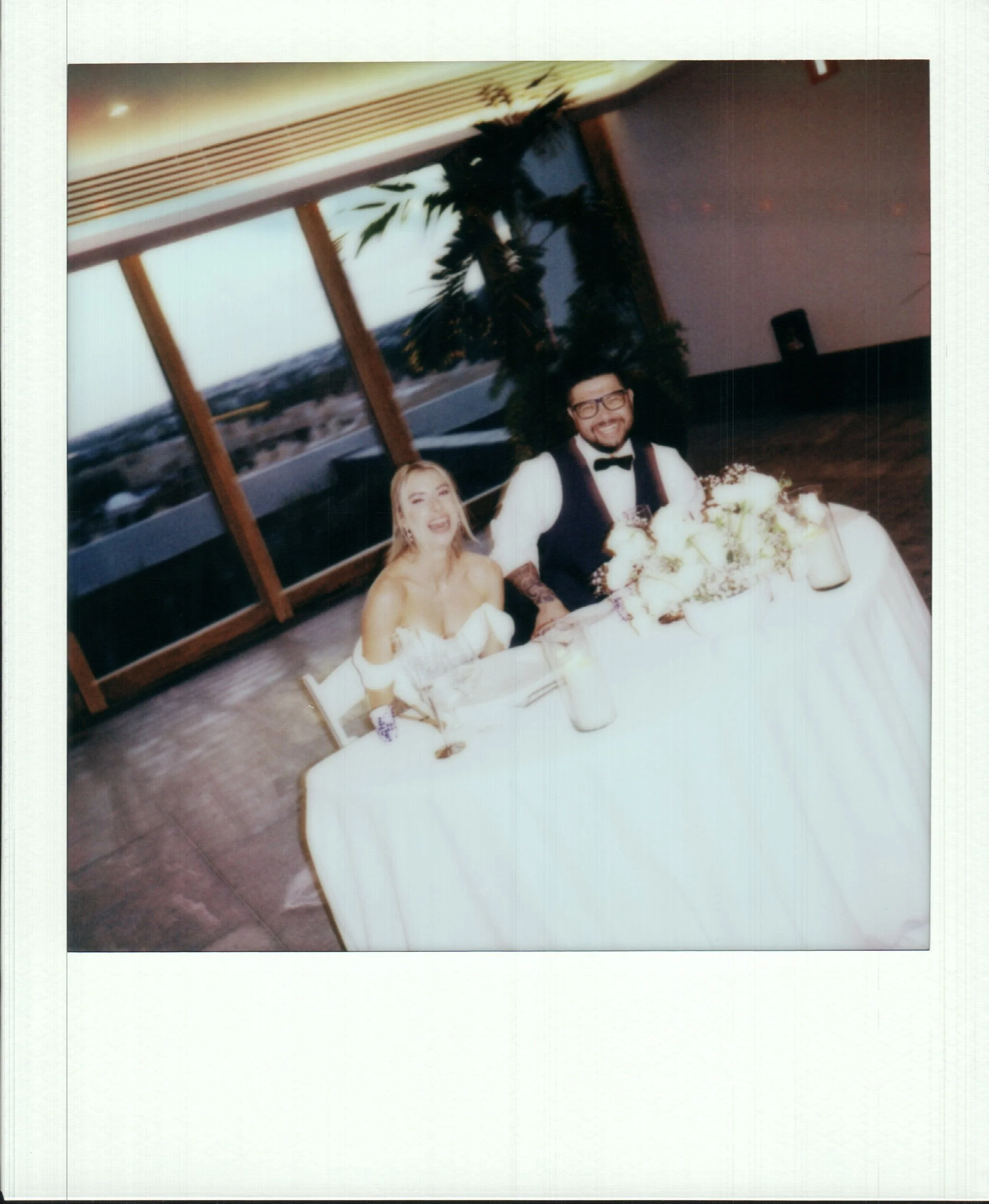 A photograph of a wedding reception with a bride and groom sitting at a table with a large floral arrangement, a woman in a white dress, and a man in a tuxedo, with decorated tables and windows in the background.