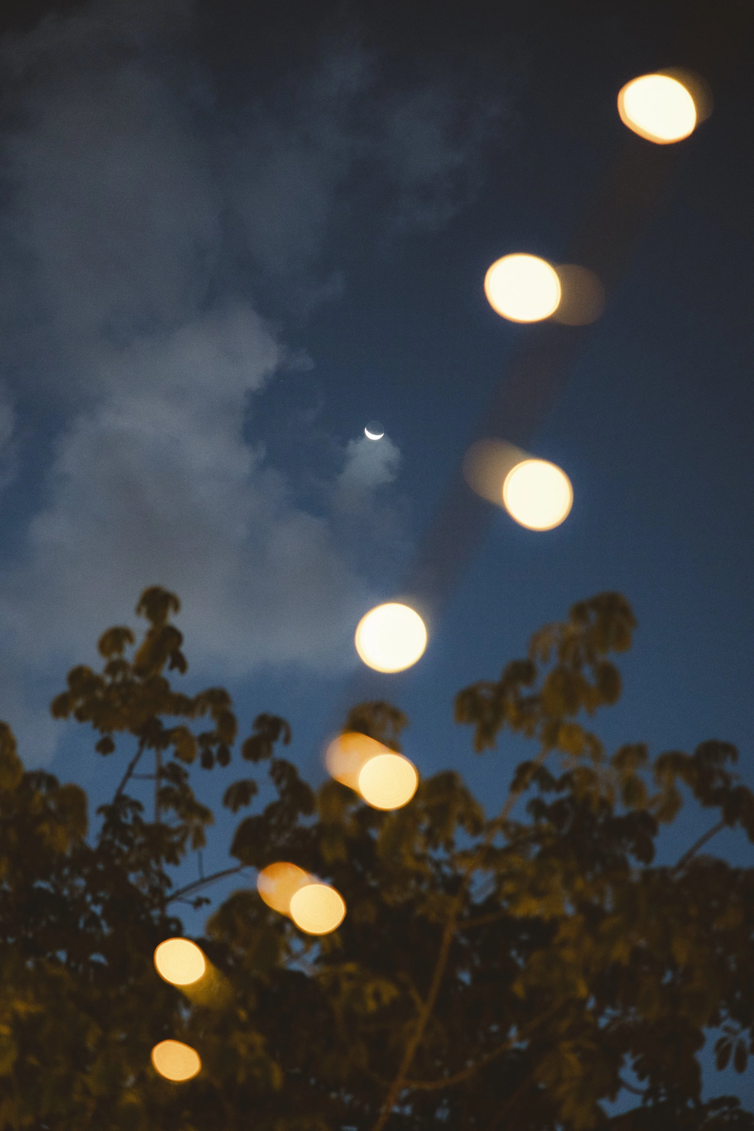 Night sky with crescent moon, clouds, and out-of-focus string lights hanging above trees.