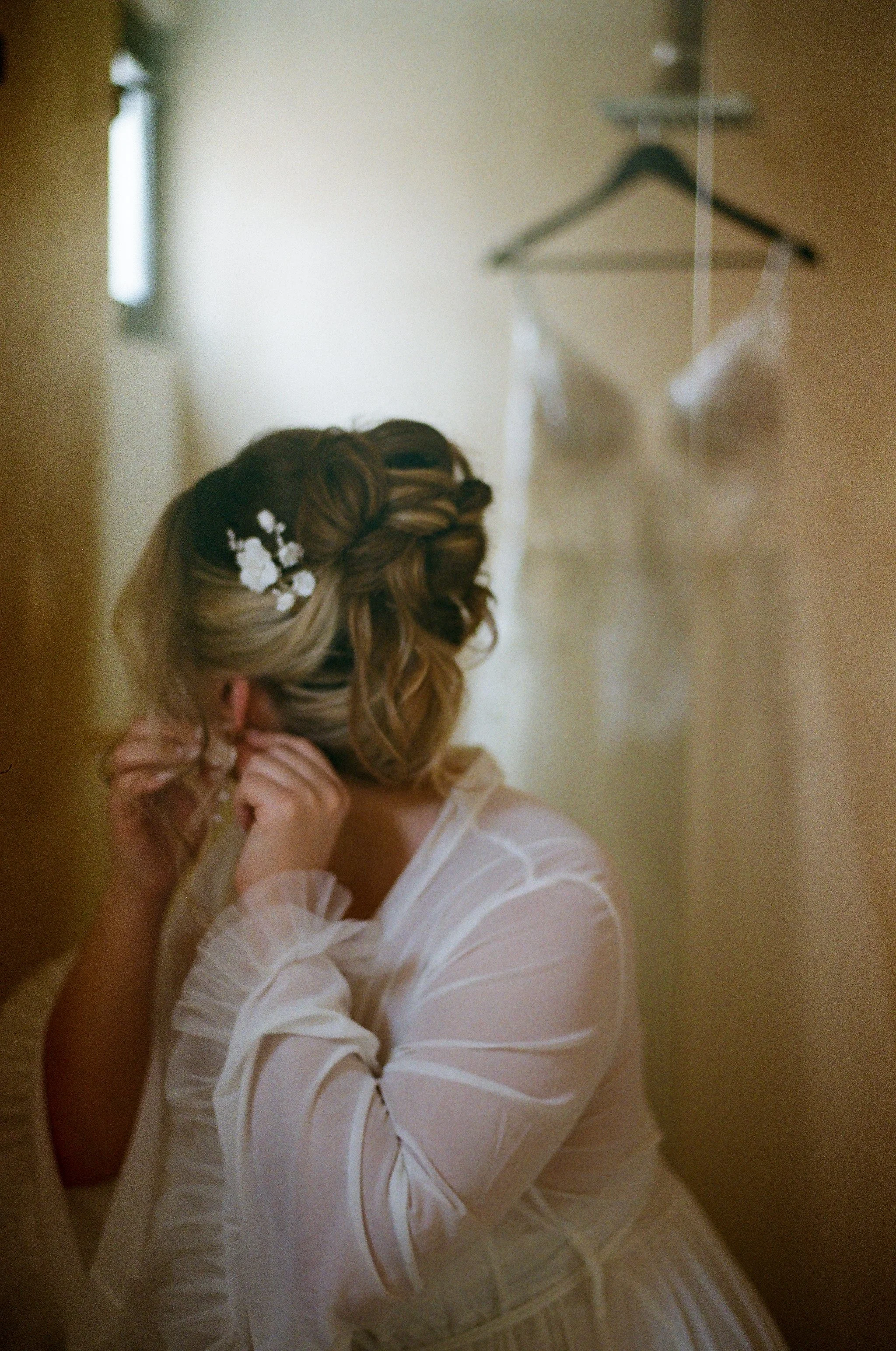 A woman with styled hair and a white dress is getting ready, touching her earring, with a hanger and dress on the wall behind her.