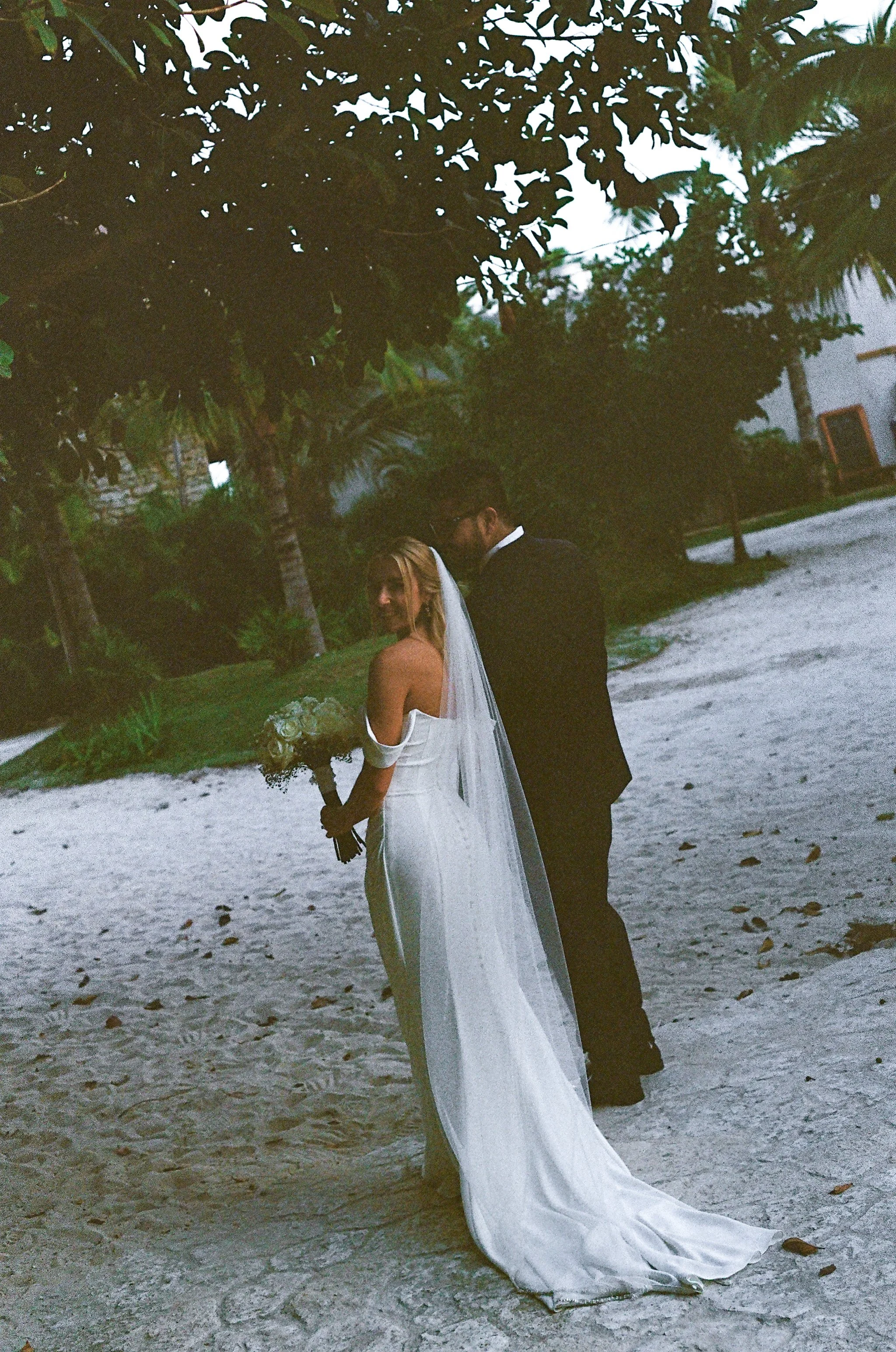 A bride in a white wedding dress holding a bouquet, standing on a sandy beach with a groom in a black suit, surrounded by tropical trees and foliage during sunset or dusk.