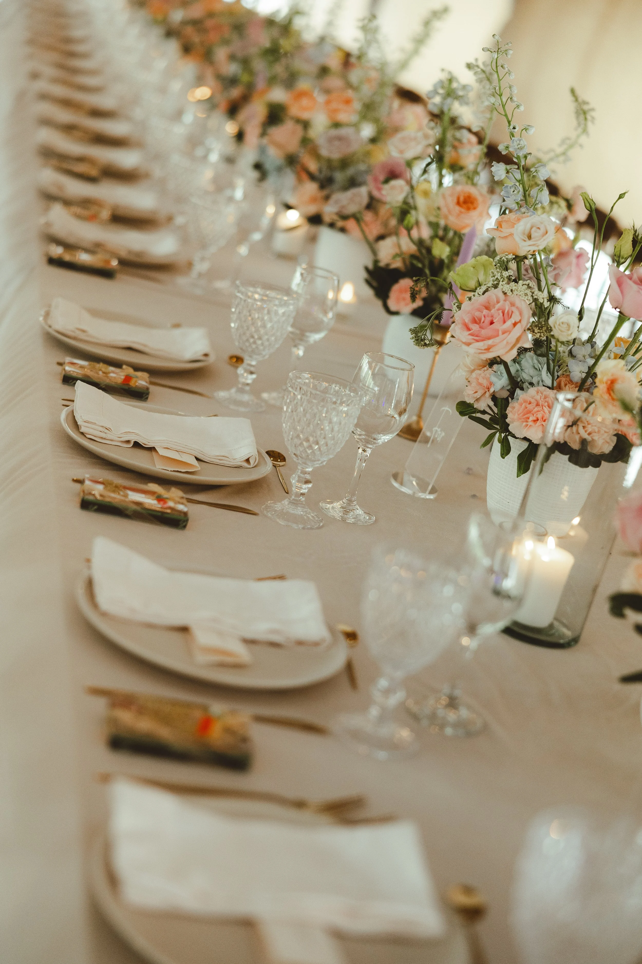 Elegant dining table decorated with pink and white roses in glass vases, surrounded by crystal glasses, white napkins, gold flatware, and lit candles for a formal event.