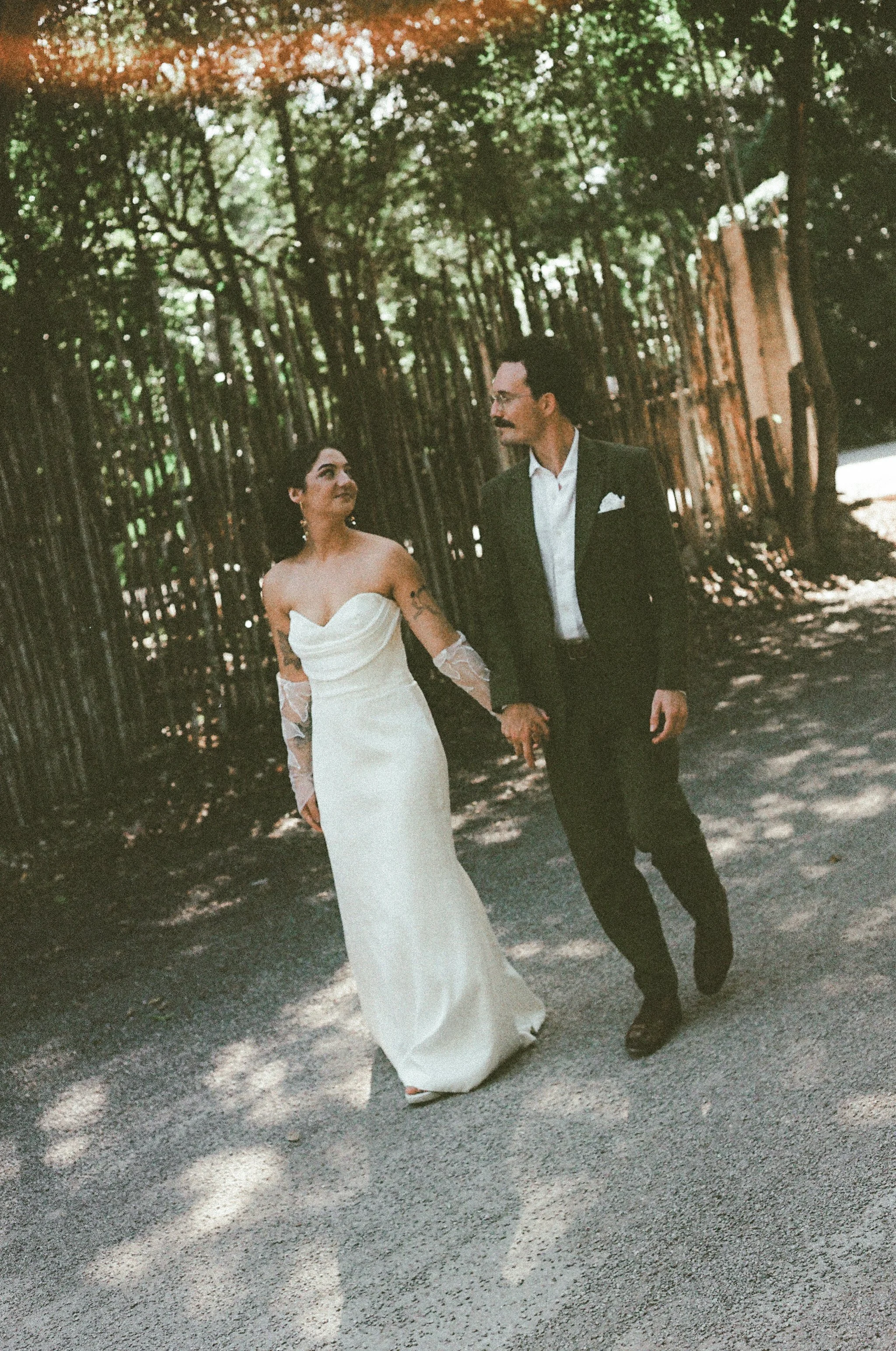 A newlywed couple holding hands and walking outdoors on a shaded gravel path in tulum. The bride is in a white strapless gown with lace sleeves, and the groom is in a dark green suit with a white shirt and pocket square.