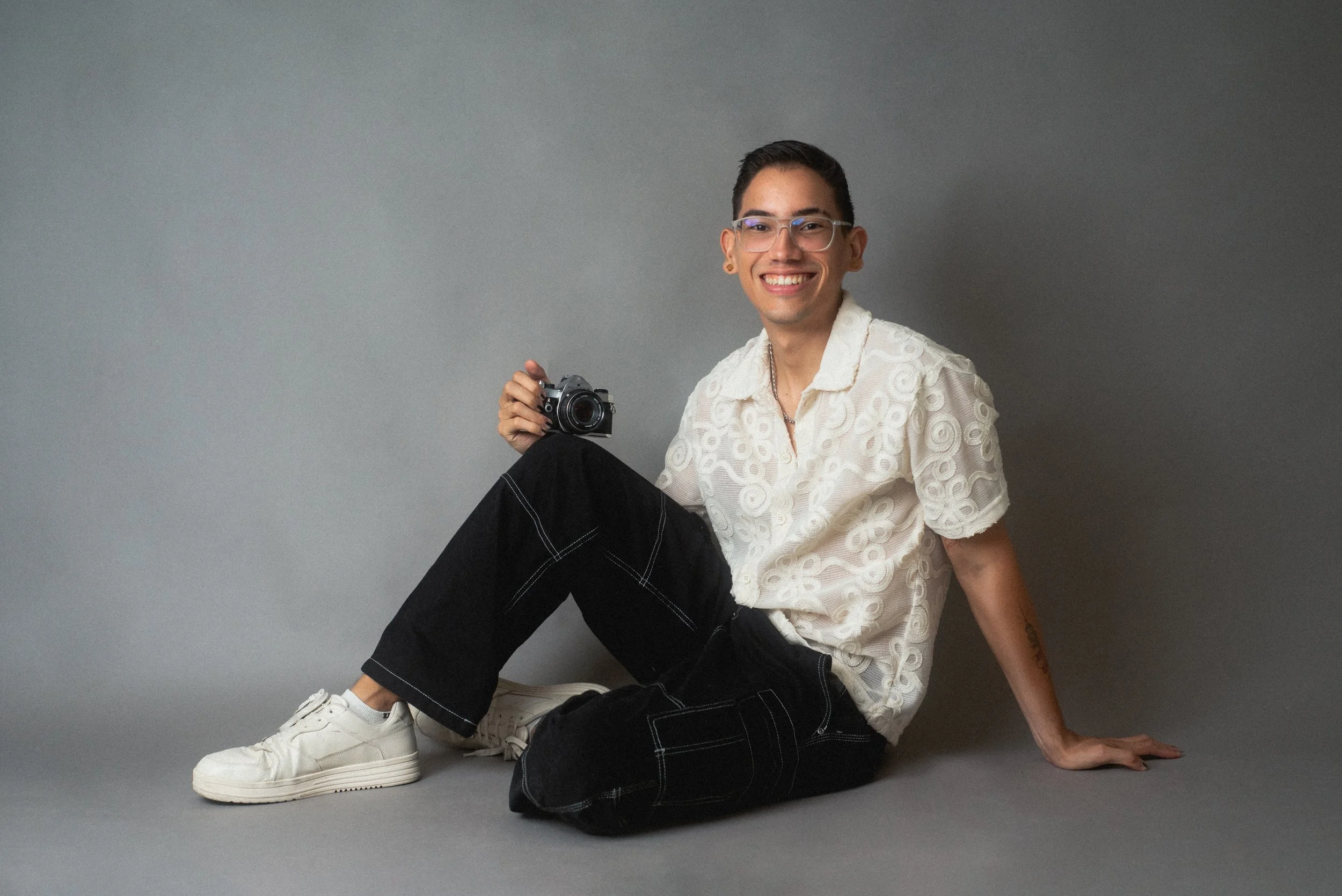 A person sitting on the floor against a gray background, smiling and holding a camera. They are wearing glasses, a white embroidered shirt, black pants with white stitching, and white sneakers. Eduardo Paralizabal wedding photography
