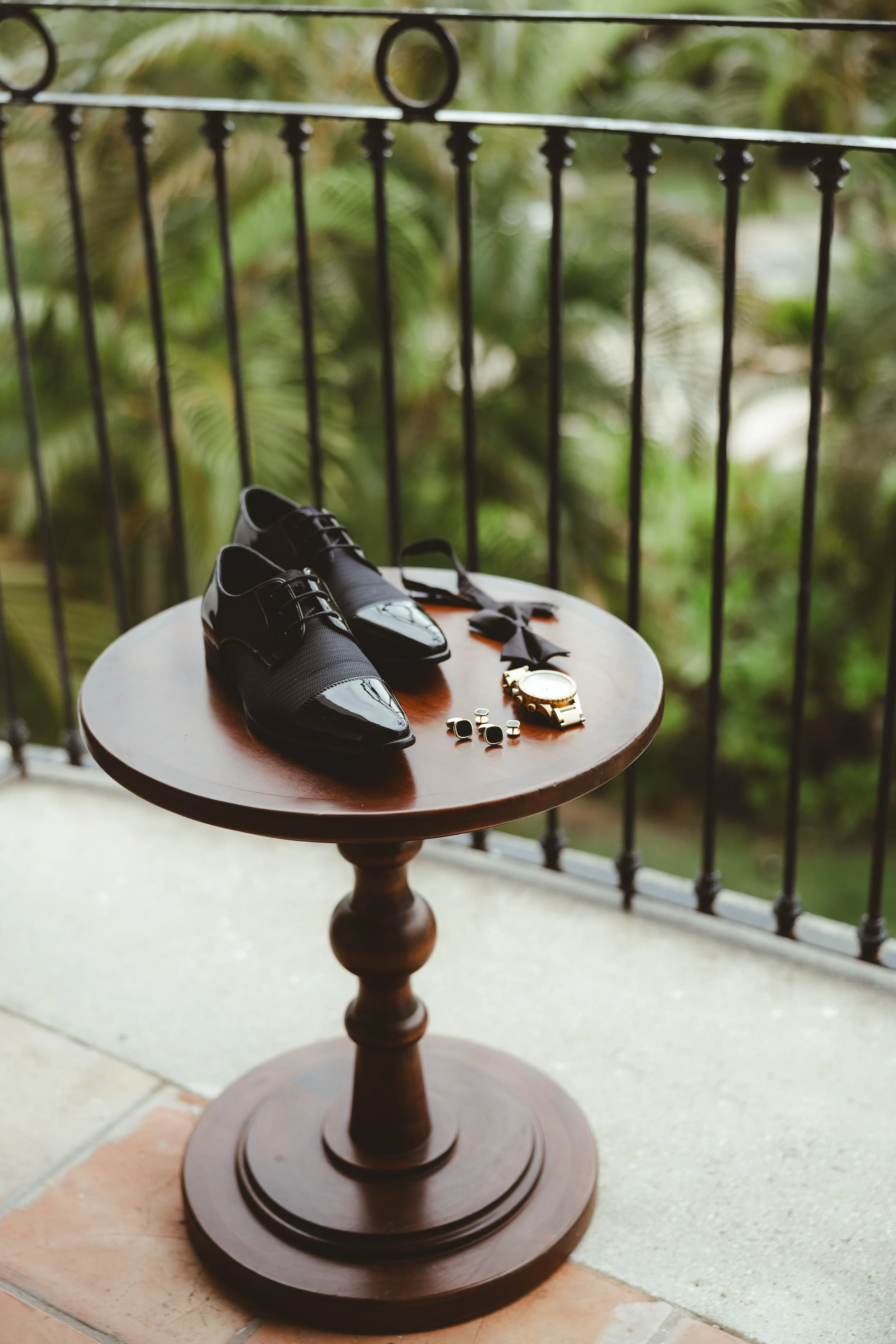 Black men's dress shoes, a watch, cufflinks, and a bow tie arranged on a round wooden table on a balcony with greenery in the background.