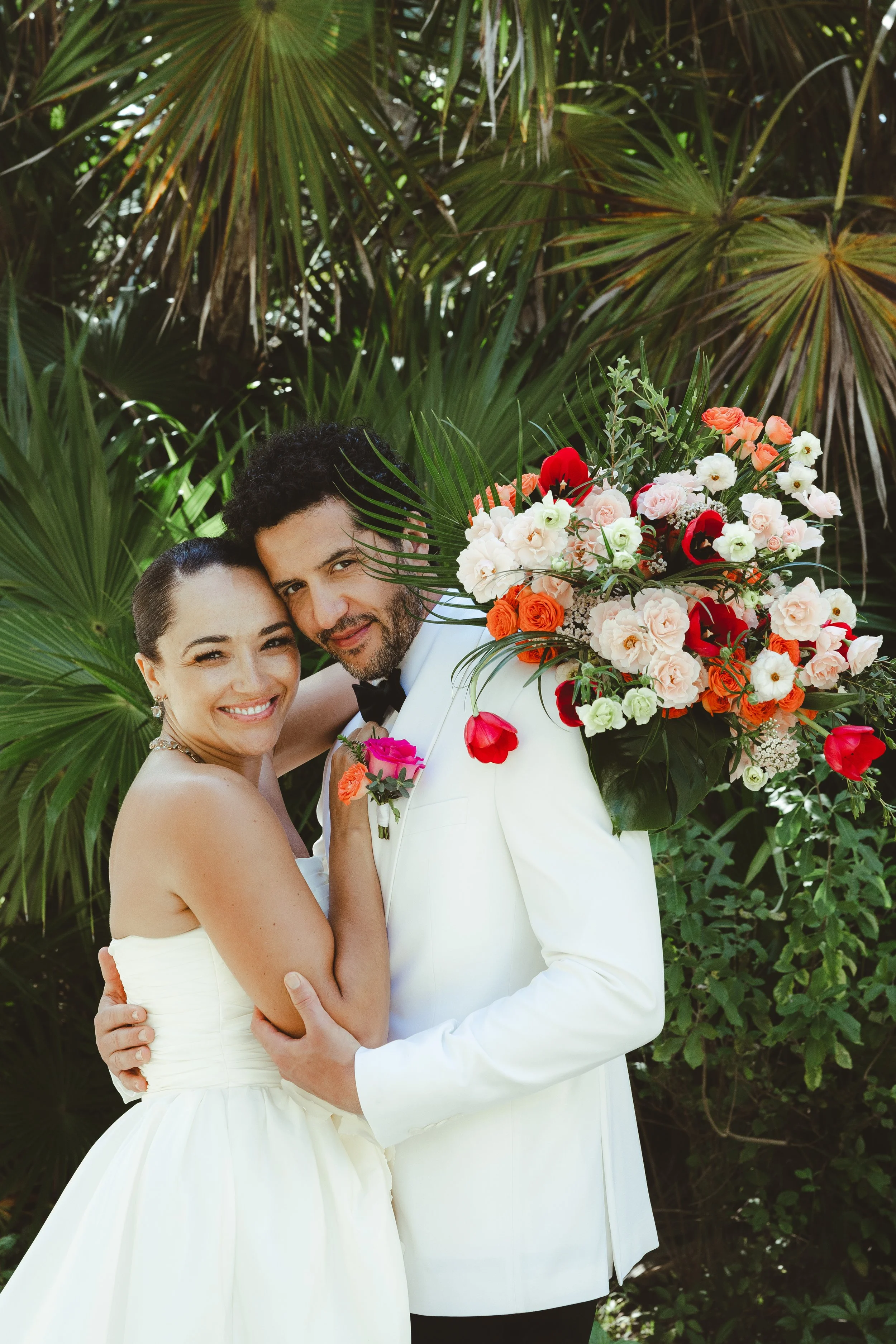 A newlywed couple smiling and embracing outdoors, with tropical plants in the background. The groom is holding a large colorful bouquet of flowers over his shoulder.