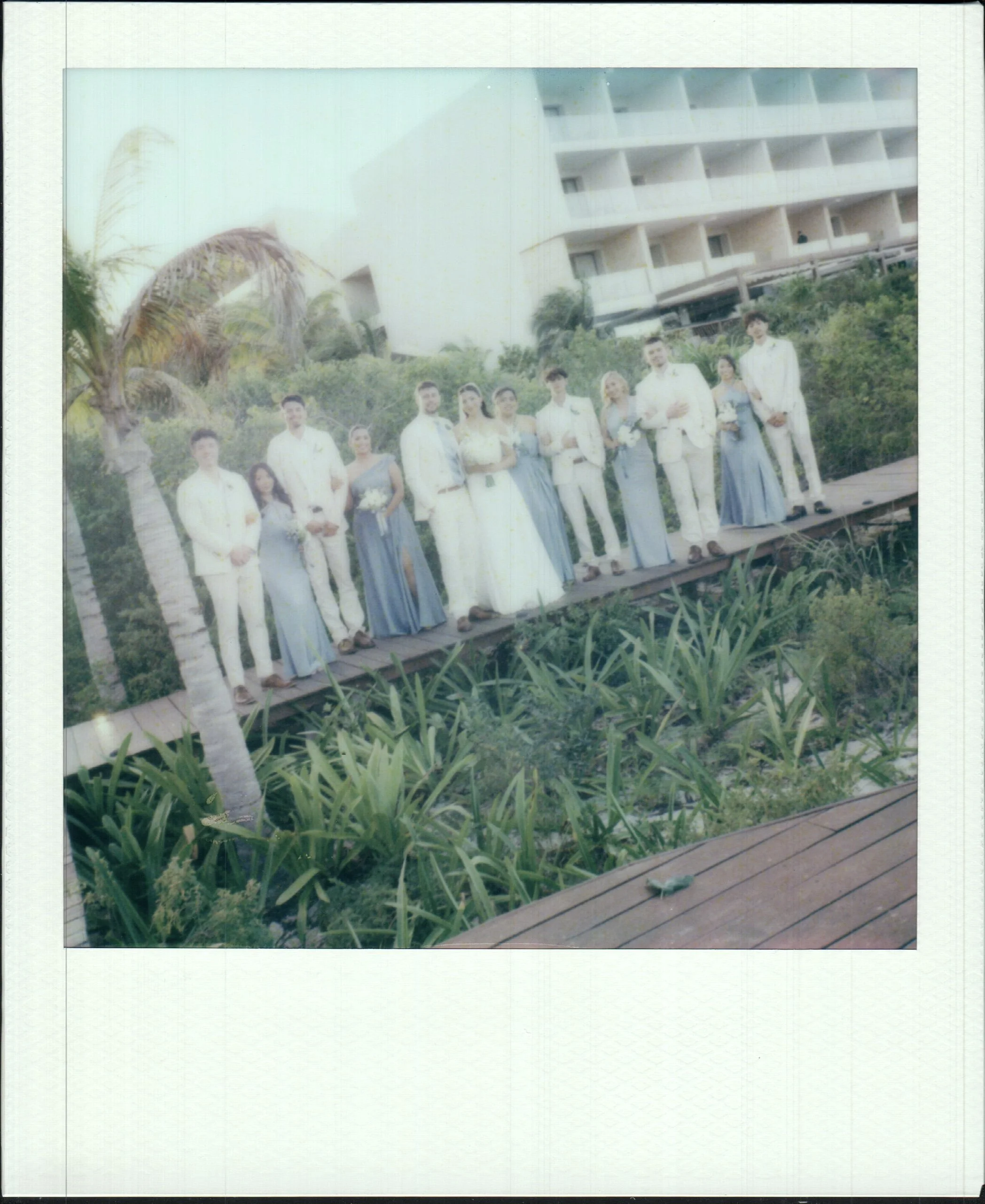 wedding party standing on a wooden platform outdoors with lush greenery and a hotel building in the background. polaroid photo.