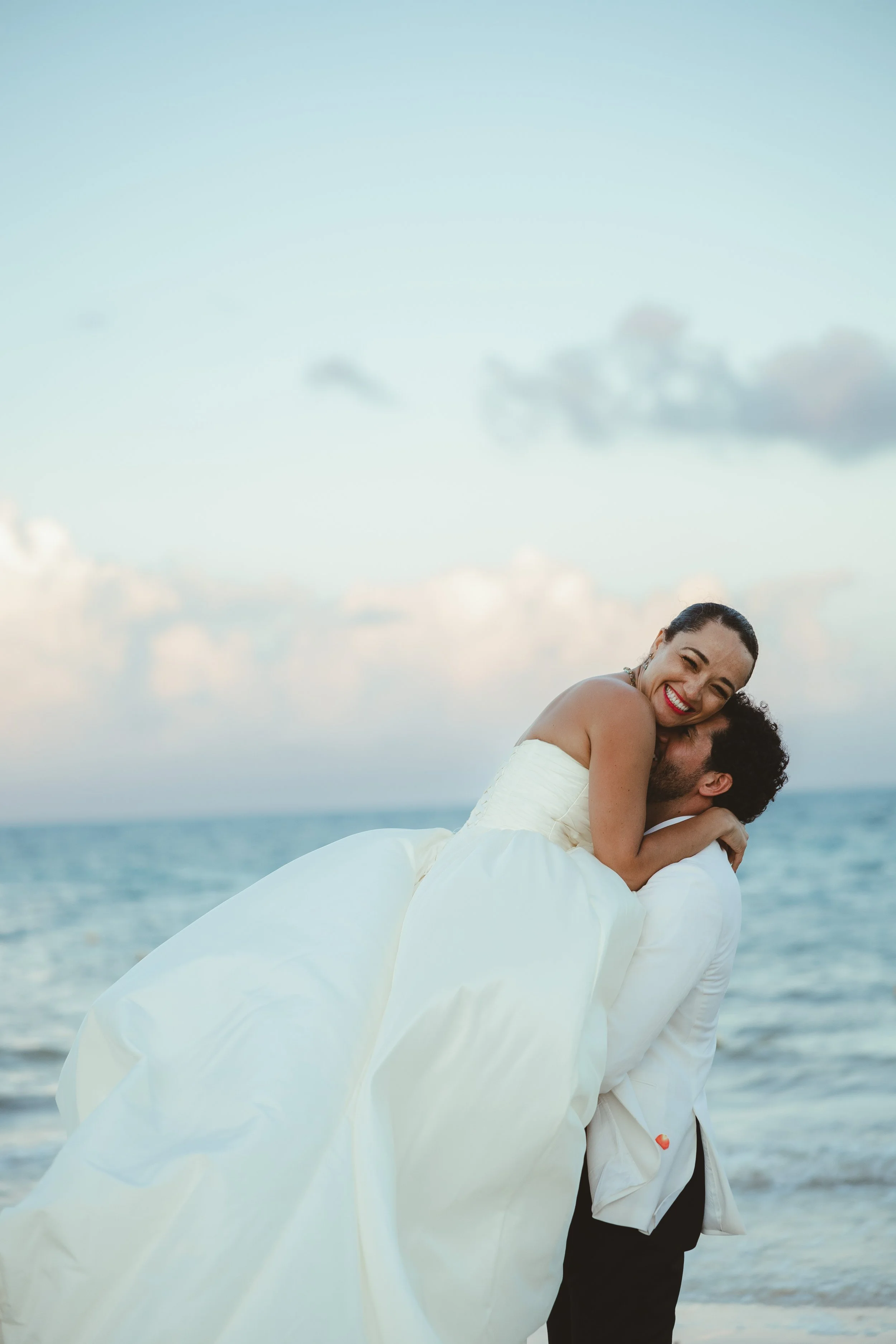 A couple in wedding attire on the beach, with the woman smiling and hugging the man while he lifts her. The woman is wearing a white wedding dress and the man is dressed in a white jacket and black pants. The ocean and sky are in the background.