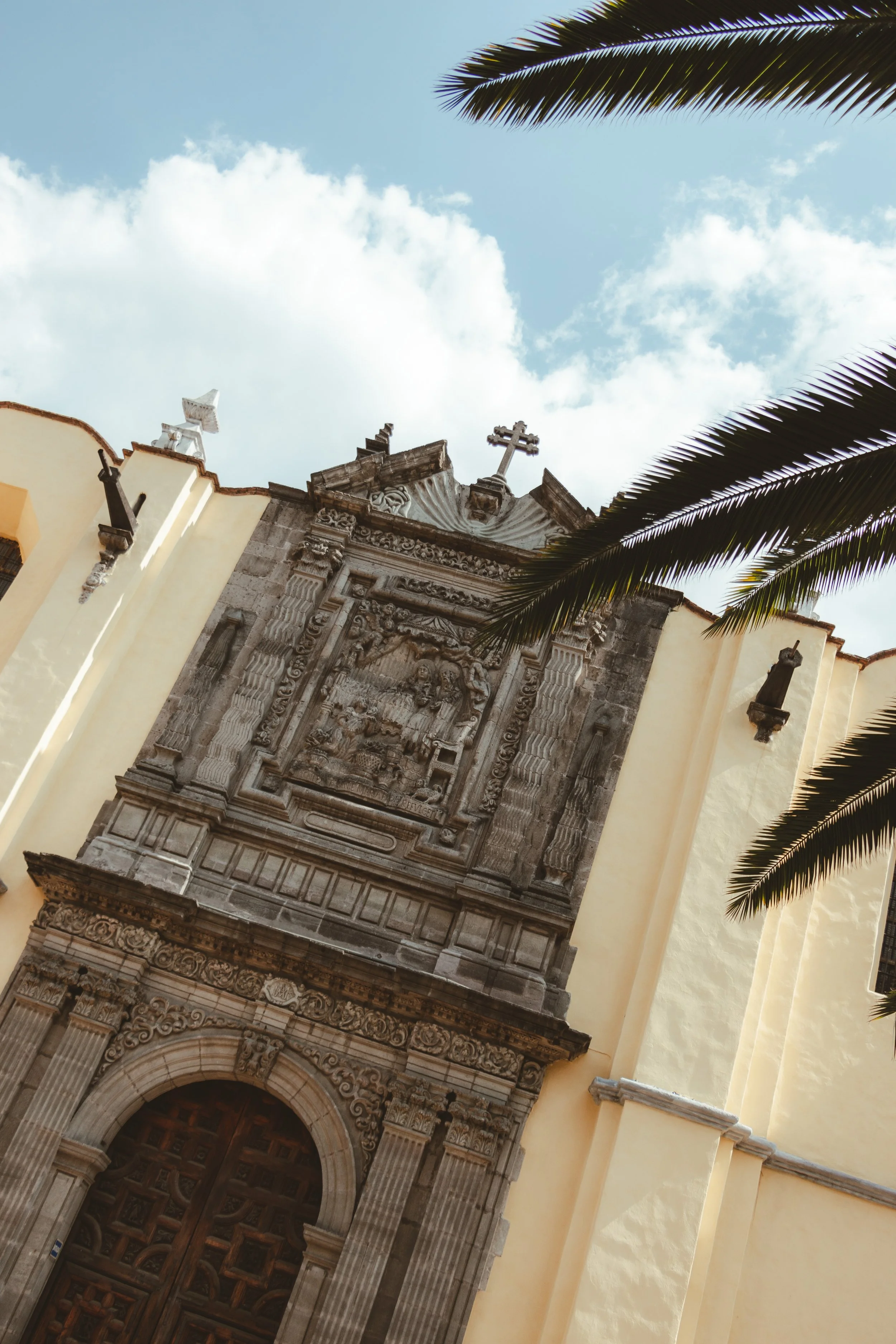A church facade with intricate stone carvings, topped with a cross, framed by palm trees against a blue sky with clouds.
