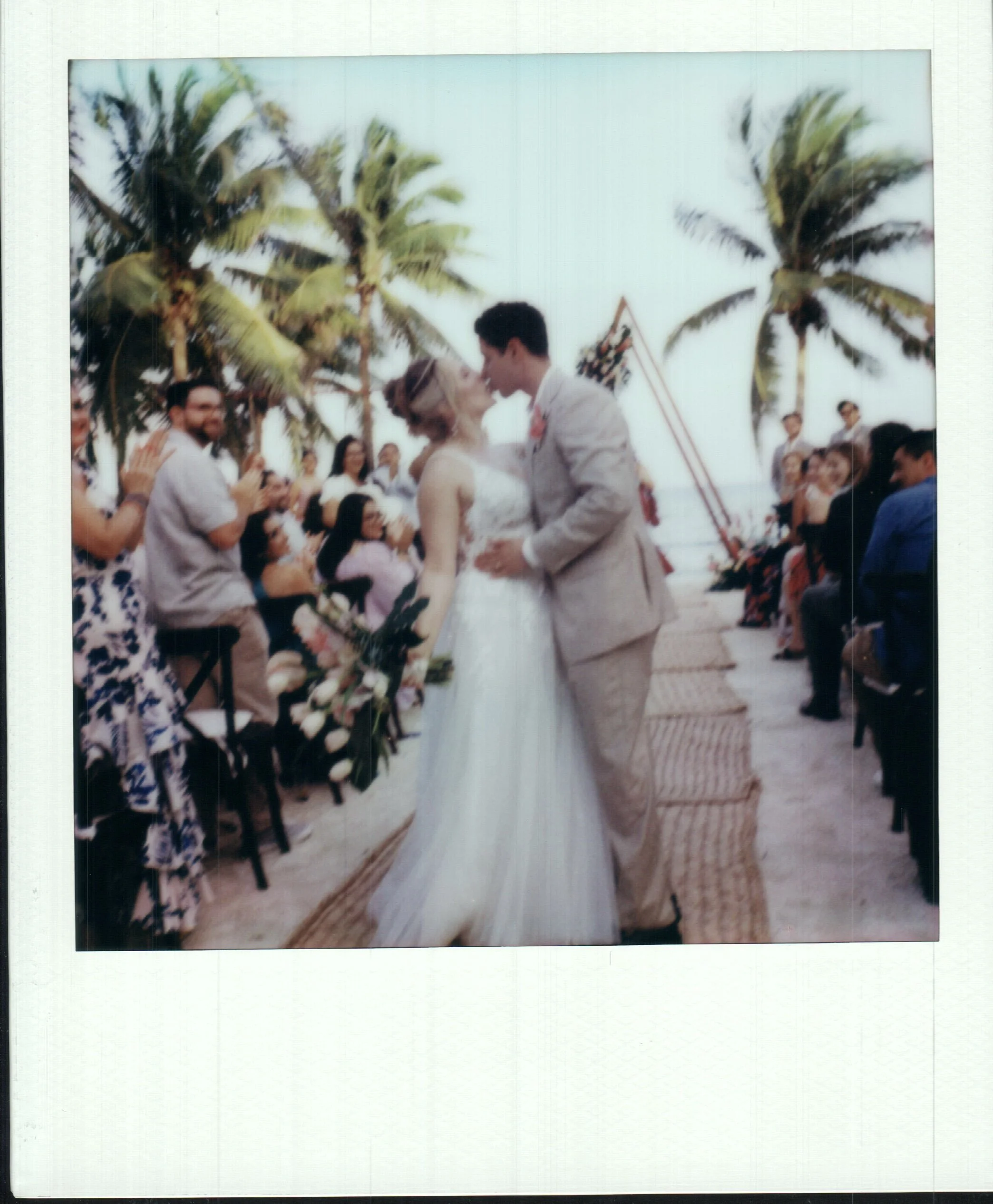 A wedding ceremony on the beach with a bride and groom kissing, surrounded by guests and palm trees, during sunset.