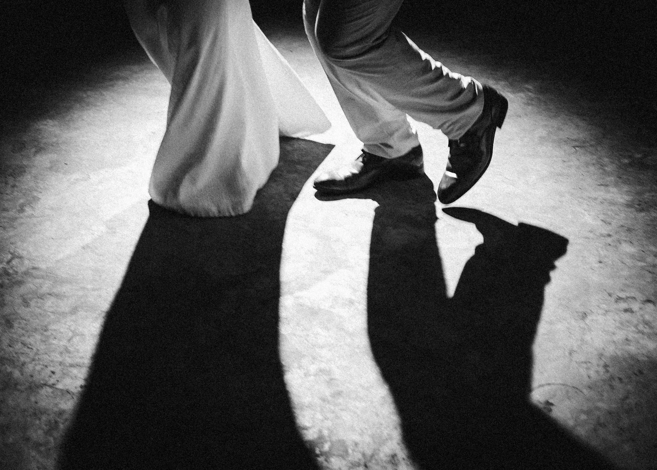 Black and white detail photograph of the shadows on the ground of a couple during their first dance, with a focus on their legs and shoes.
