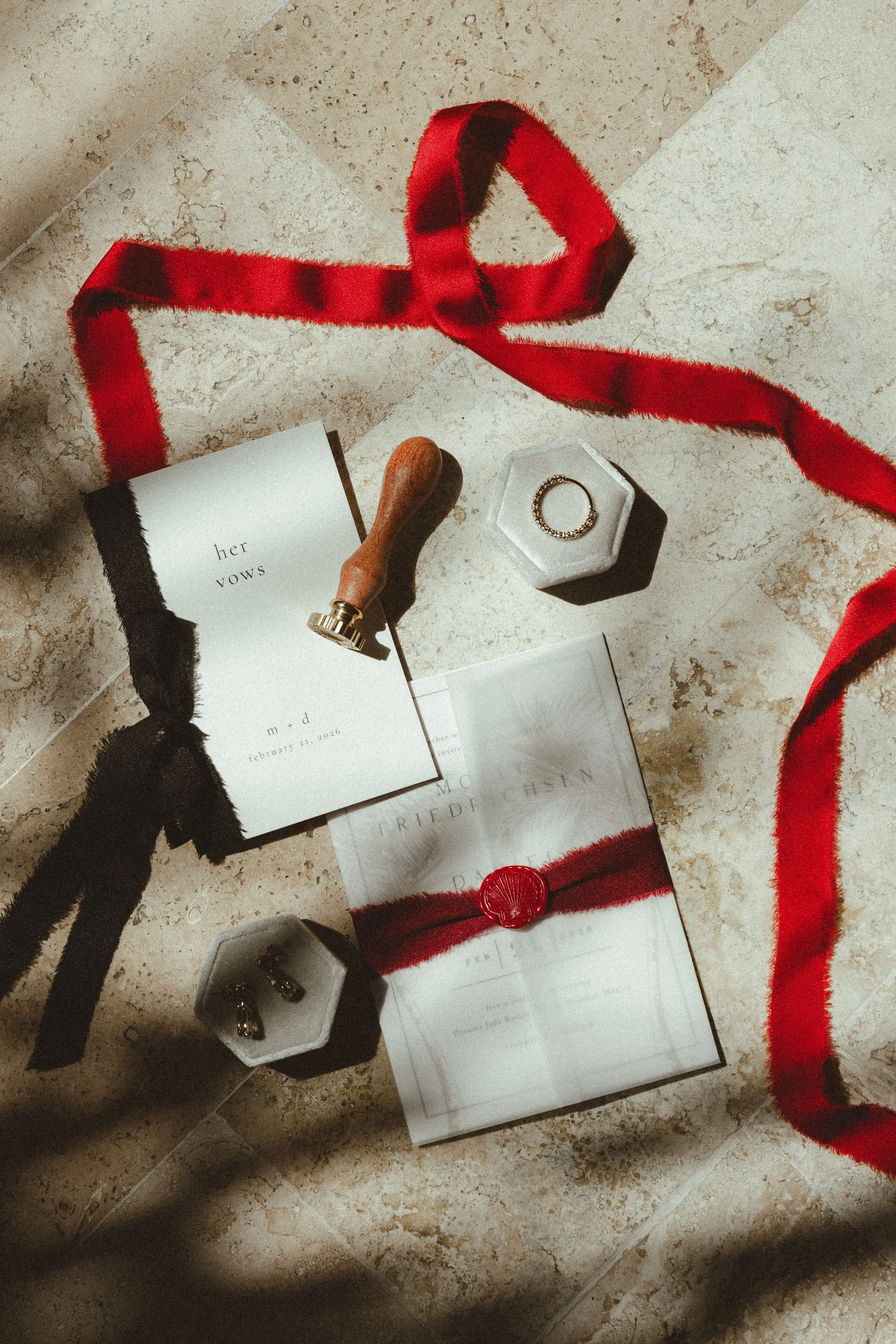 A flat lay of red wedding-related items on a marble floor, including a red and black ribbon, a ring in a hexagonal dish, a wedding vow booklet, a wax sealing stamp, and some jewelry in black and white boxes.