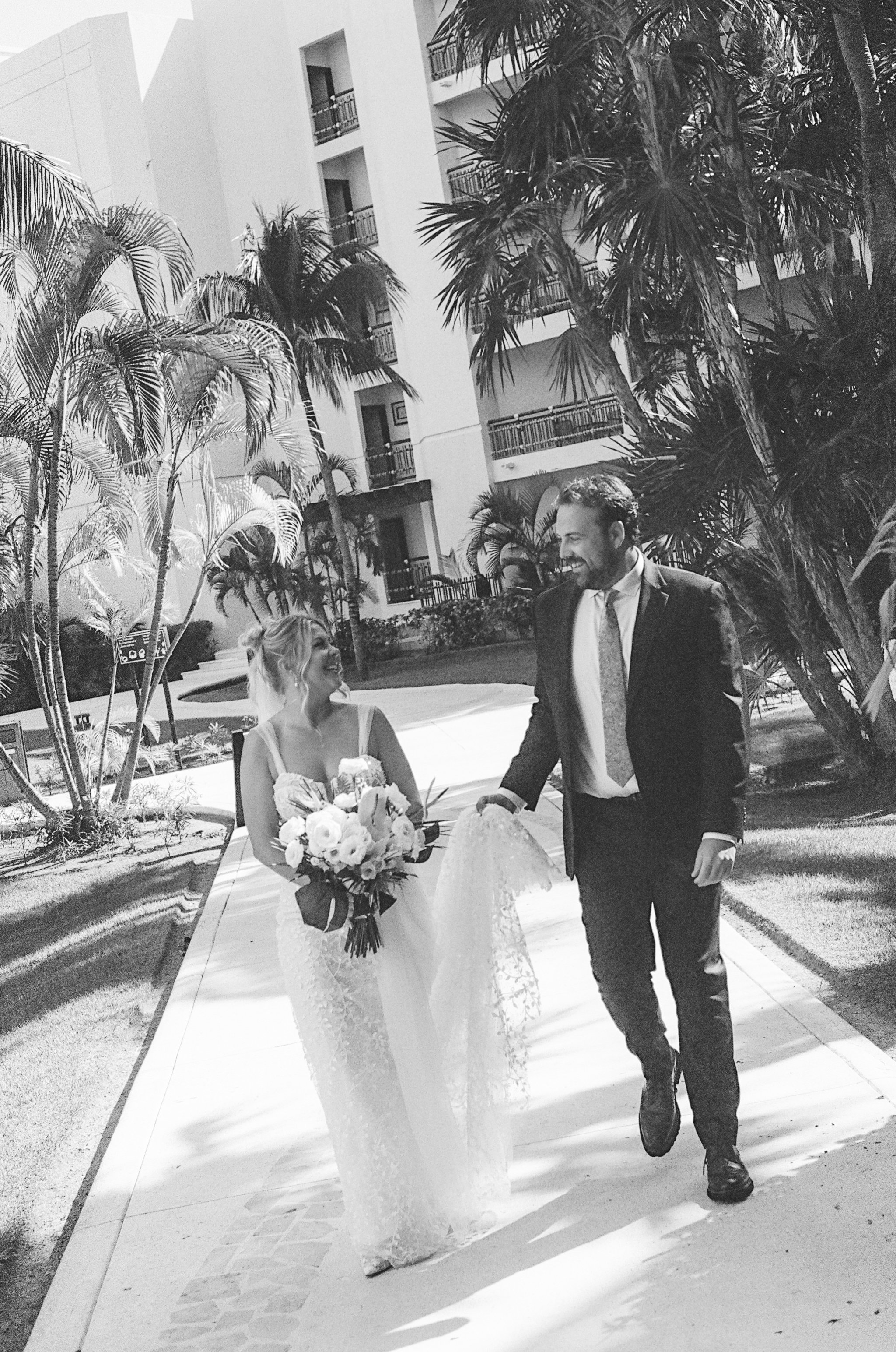A black-and-white photo of a bride and groom walking outside on a sunny day, holding hands. The bride is wearing a lace wedding dress and holding a bouquet, and the groom is in a suit. There are palm trees and a modern apartment building in the backg