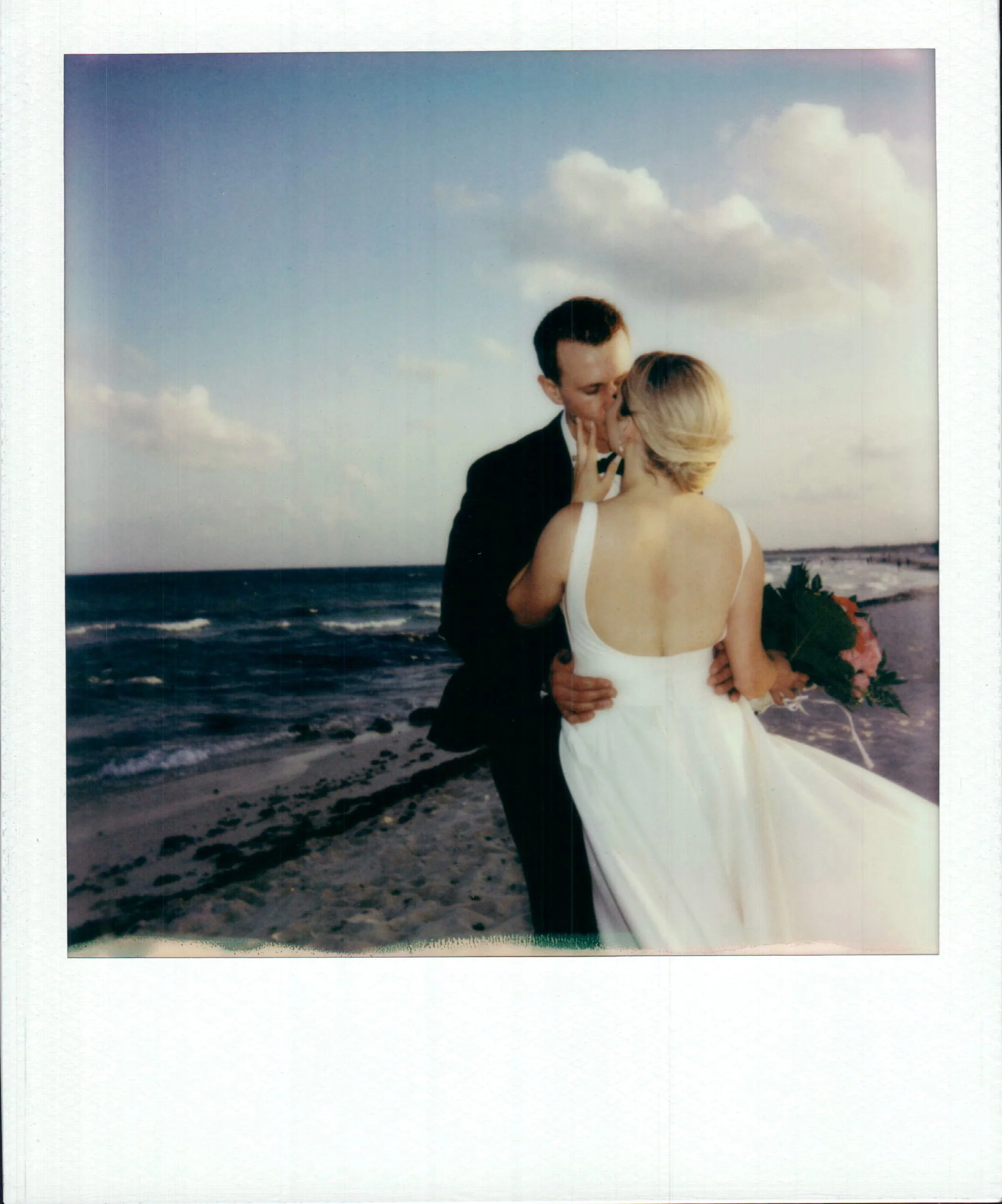 A newlywed couple sharing a kiss on the beach, with the ocean and sky in the background.