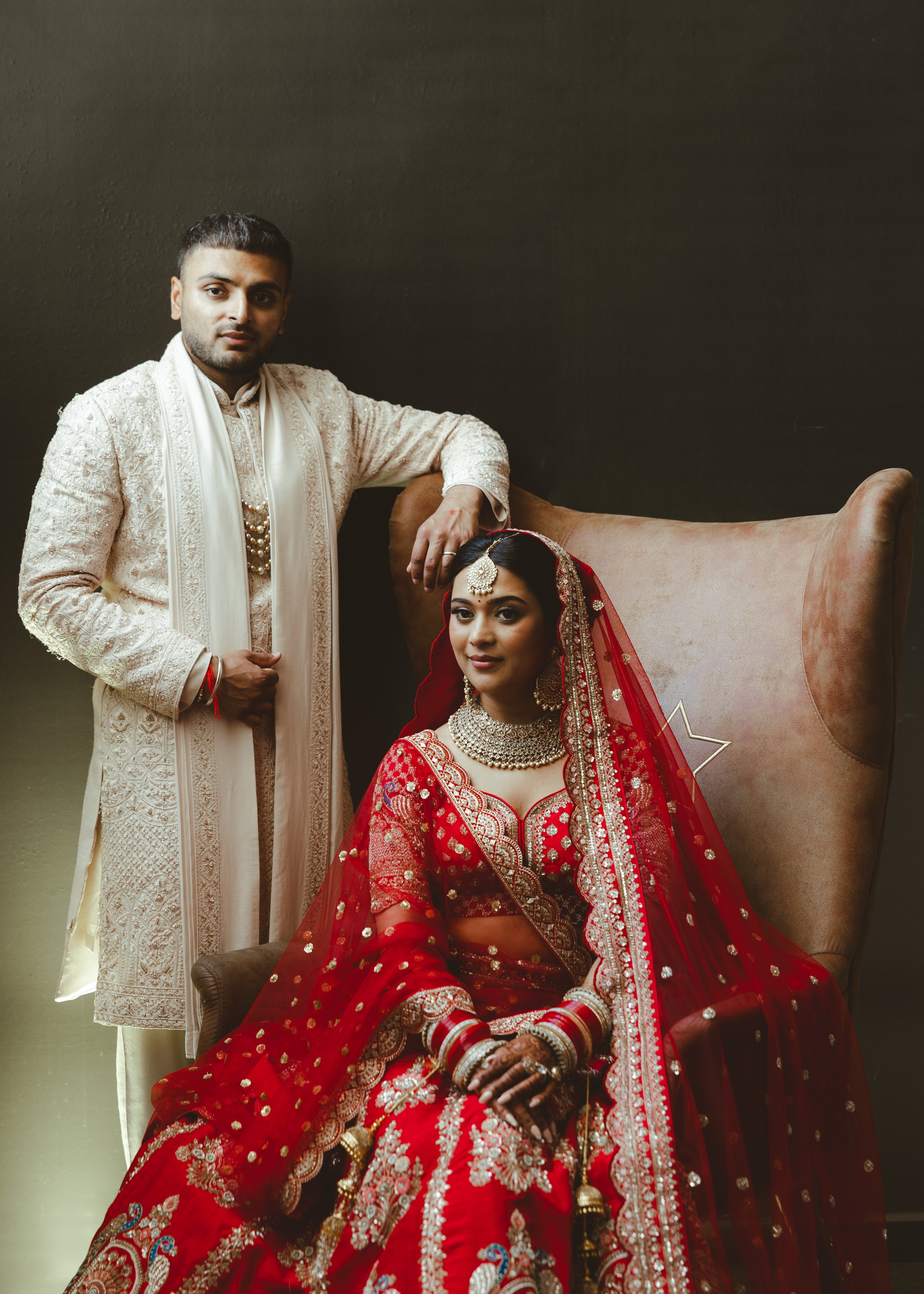 A bride dressed in a red embroidered bridal outfit with jewelry and henna sitting on a chair, and a groom in traditional cream-colored attire standing beside her, against a dark background.