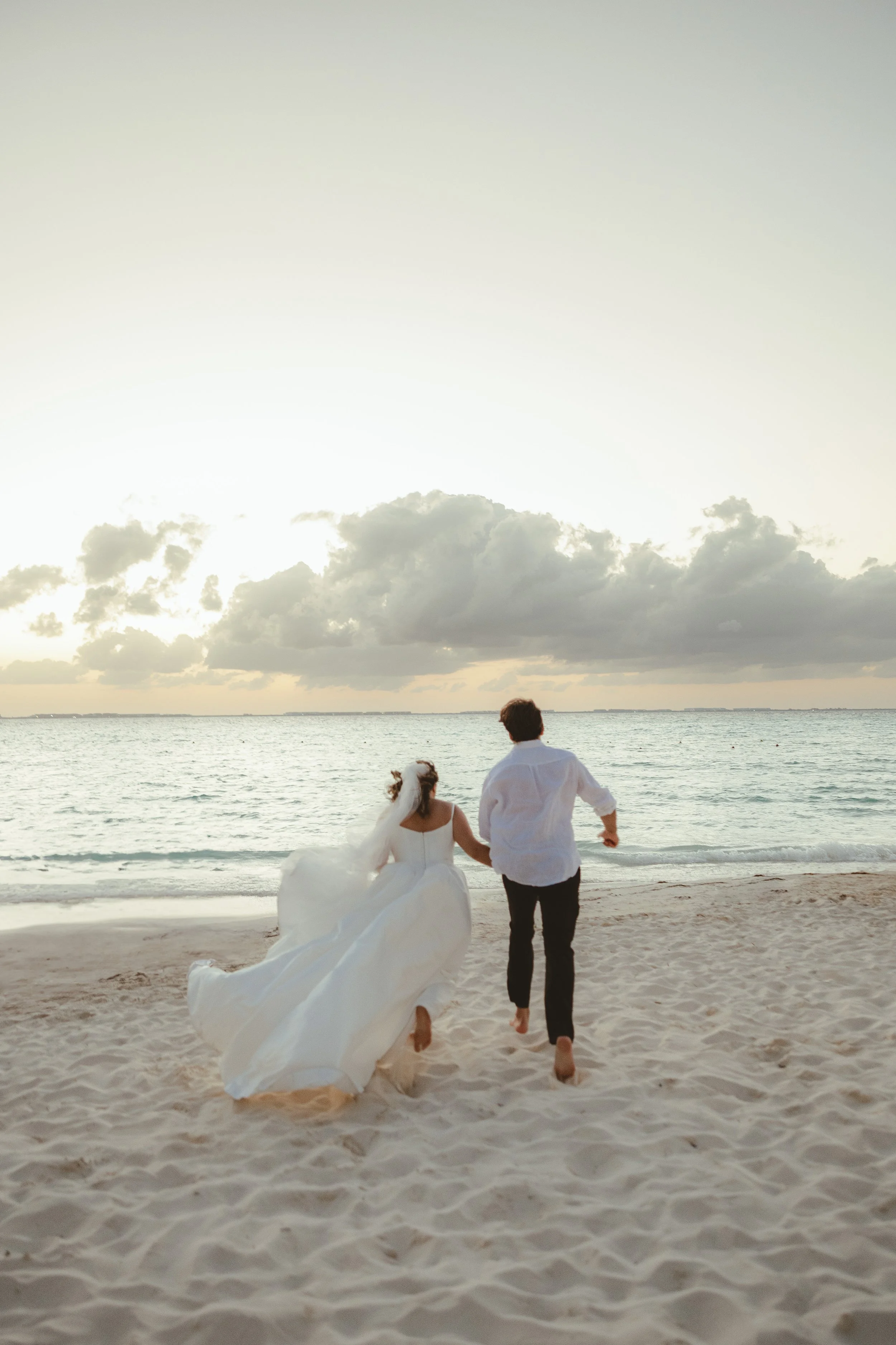 A bride and groom dressed in wedding attire are walking along the beach at sunset, holding hands, with the ocean and cloudy sky in the background.