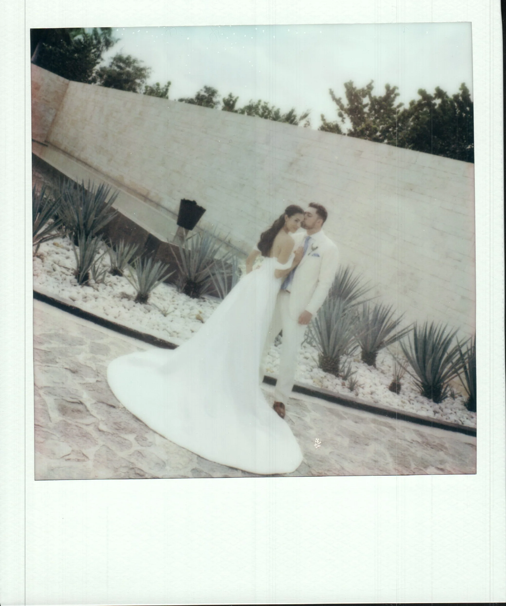 A bride and groom stand together outdoors, with the bride in a white wedding gown and the groom in a light-colored suit, against a background of a low white wall and trees.
