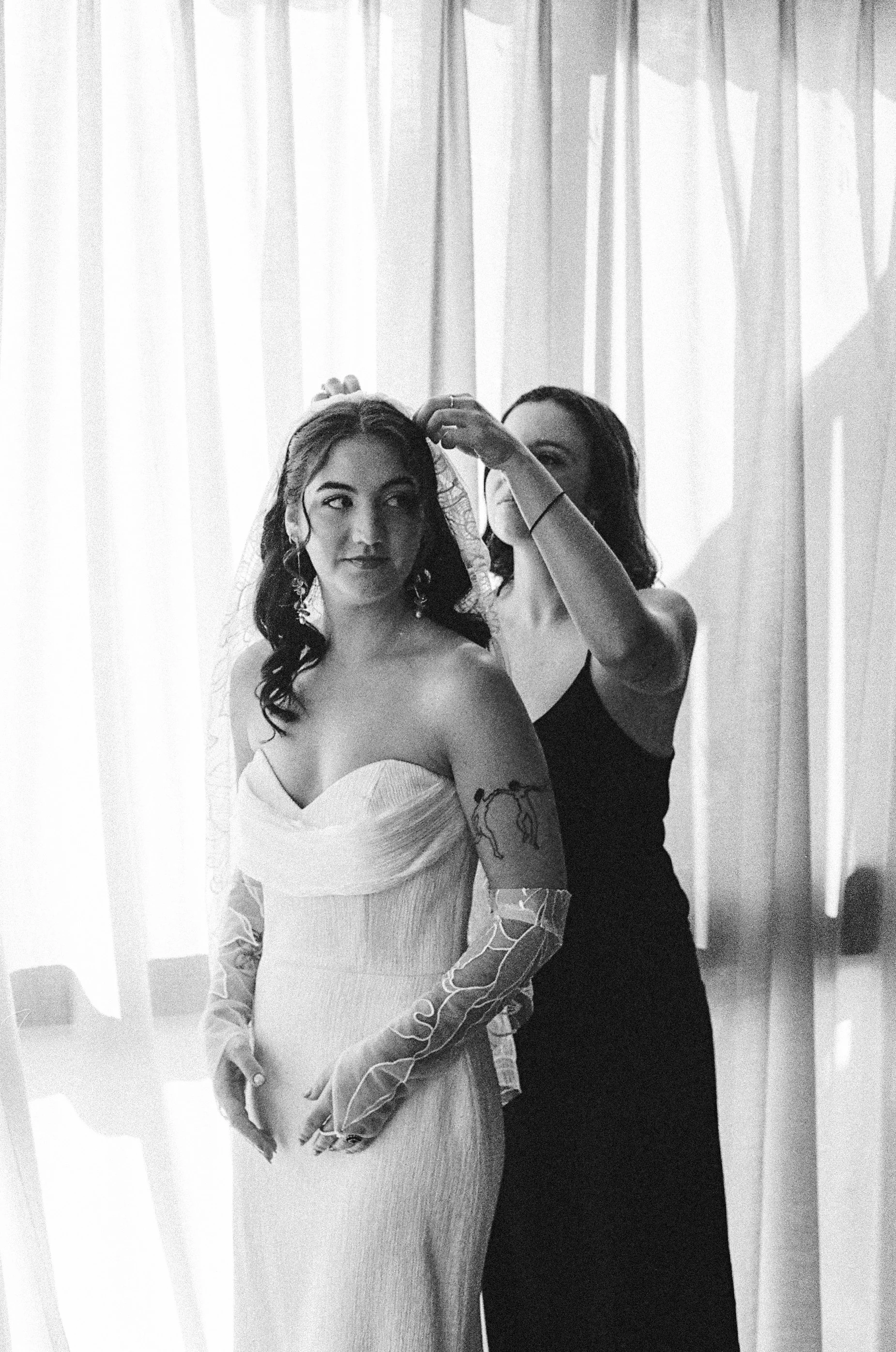 A bride in a wedding dress getting her veil adjusted by her bridesmaid indoors with curtains in the background.
