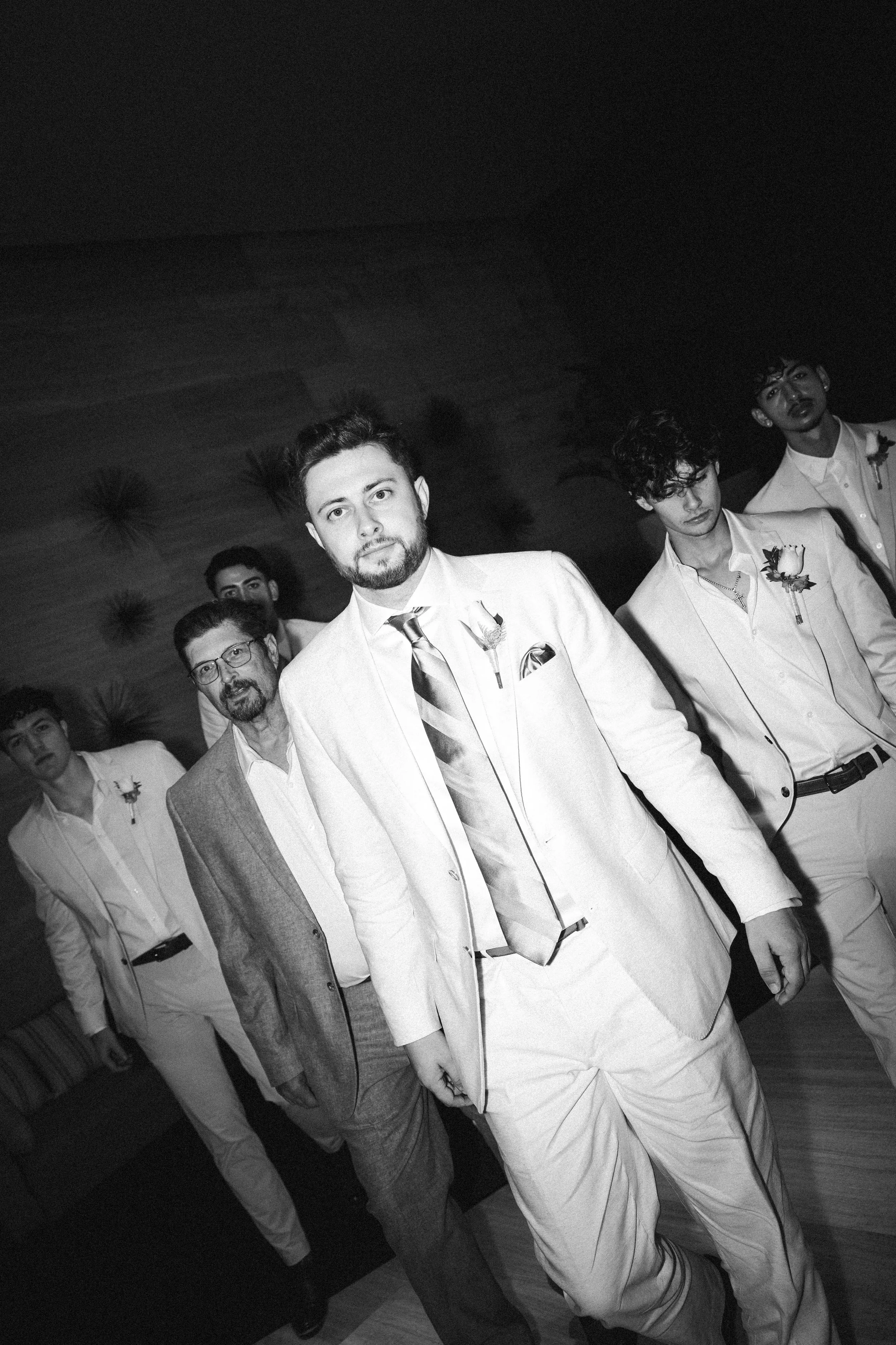 Group of groomsmen in formal attire walking together in a dimly lit indoor space.