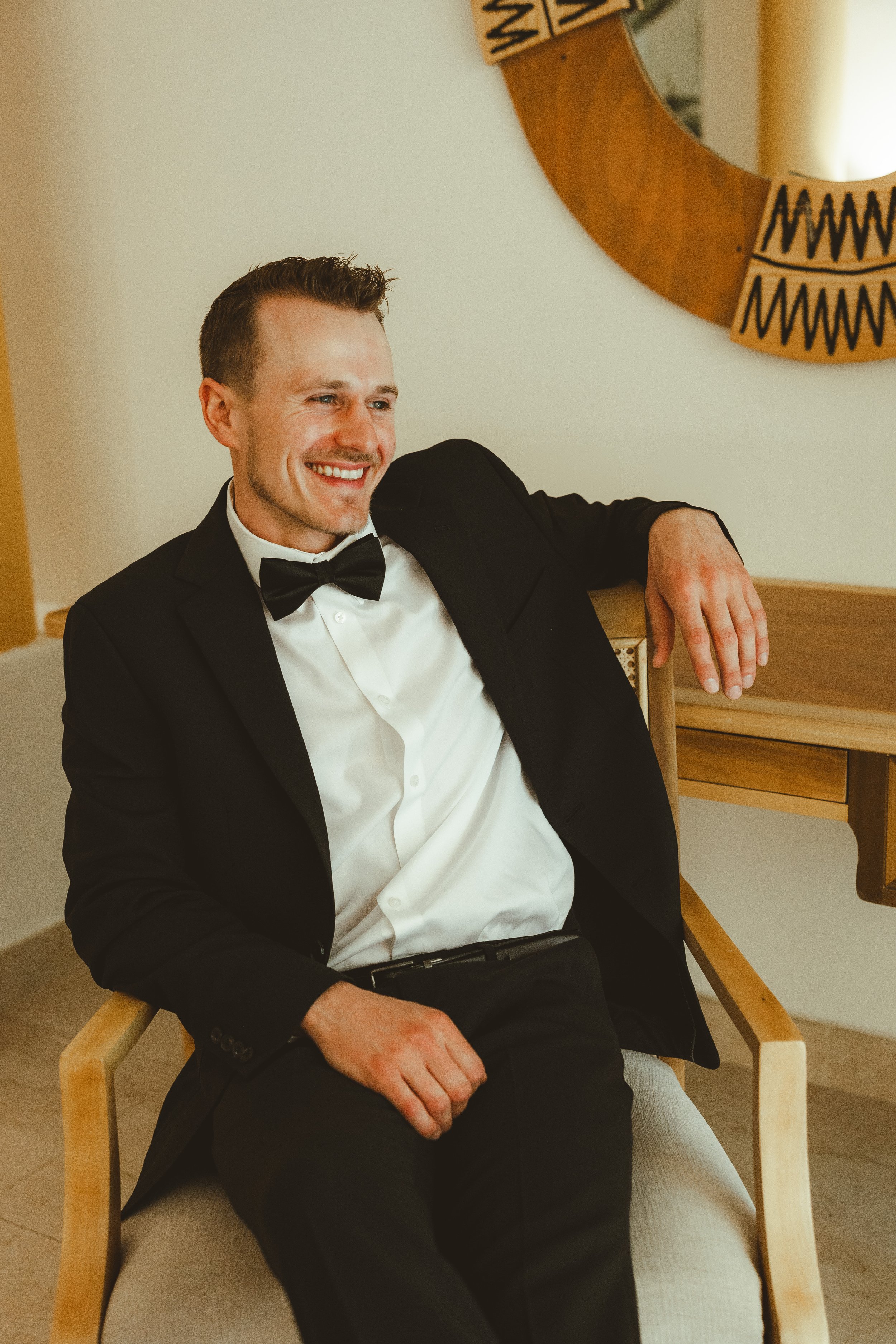 A man in a tuxedo with a black bow tie, sitting in a wooden chair, smiling, with a round mirror on the wall behind him.
