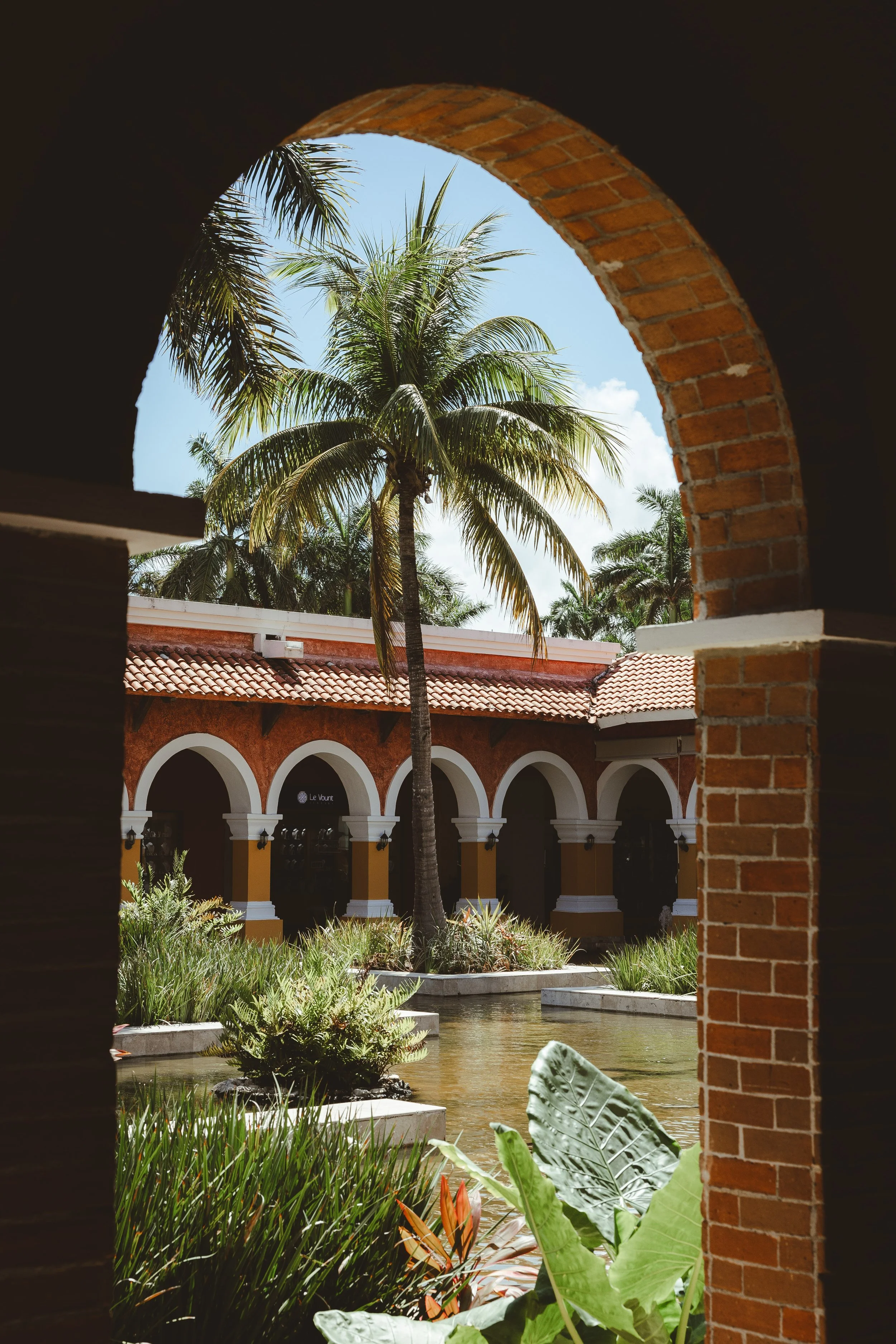 Tropical courtyard with palm trees, water feature, and terracotta building walls with arches, seen through a brick archway.