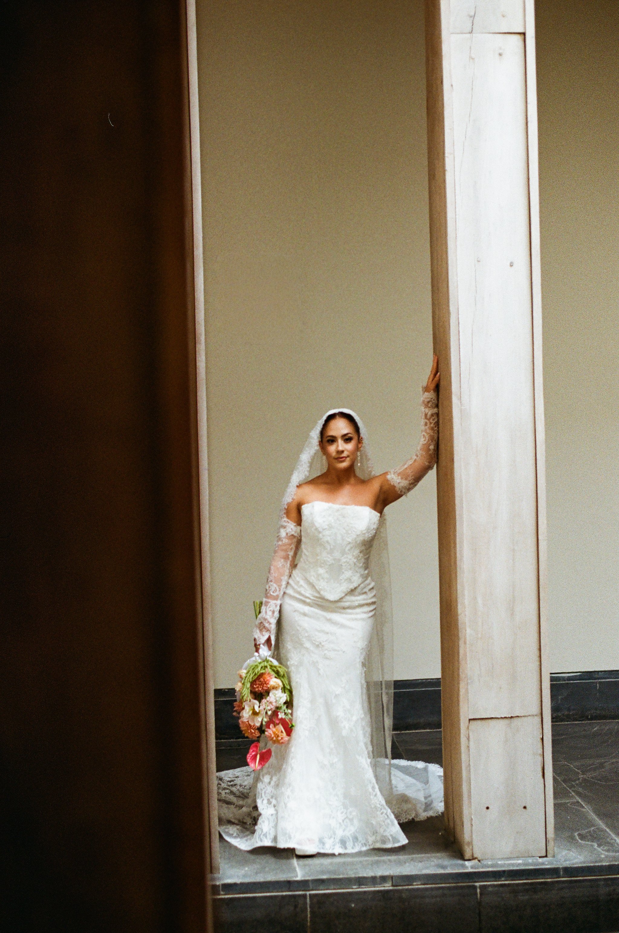 Bride in a white lace wedding gown holding a bouquet of colorful flowers, standing in front of a camera