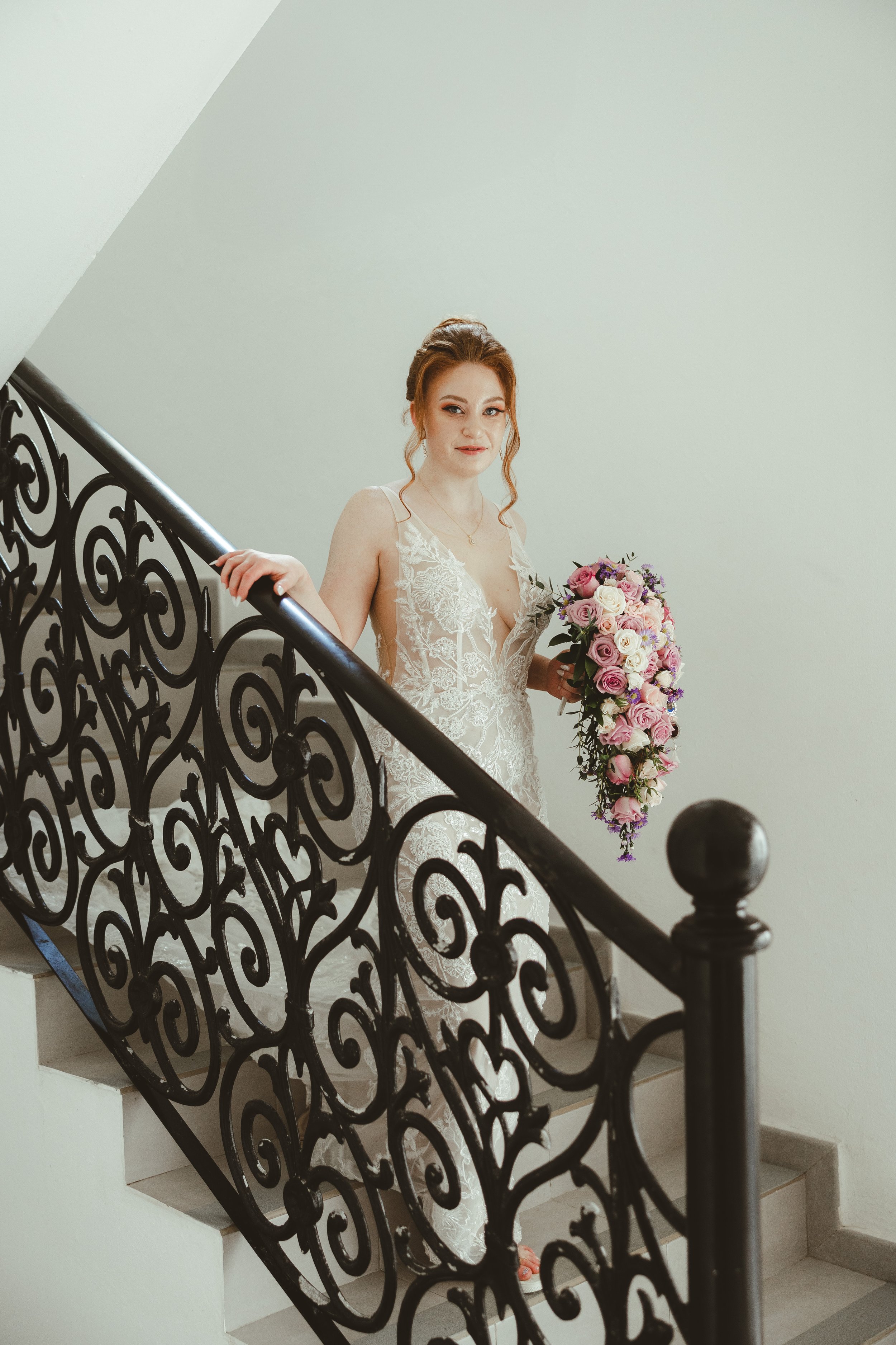 A woman in a wedding dress holding a floral bouquet standing on a staircase with an ornate black metal railing.