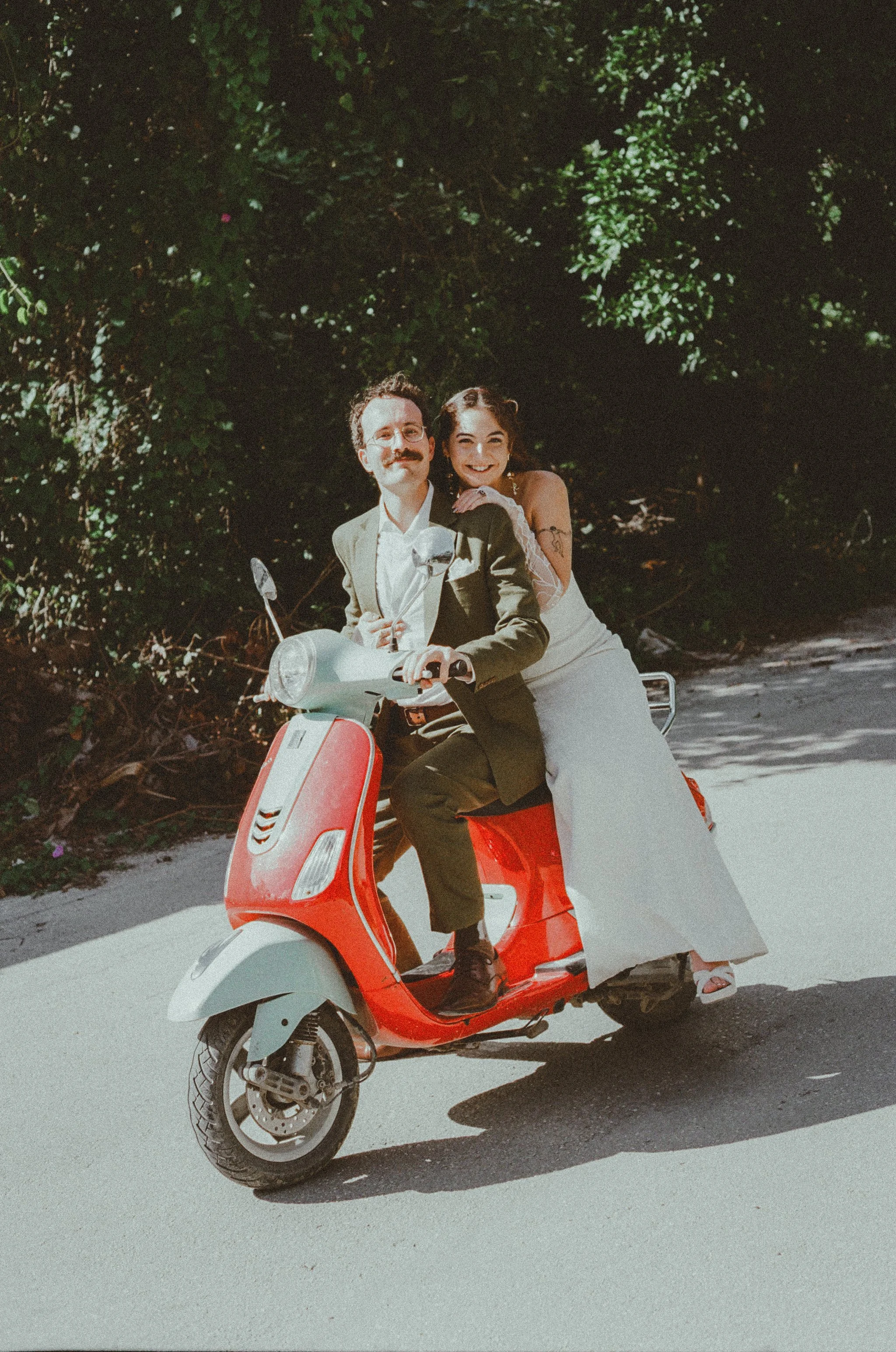 A man and woman in wedding attire riding a red and white scooter on a sunny day, surrounded by green foliage.