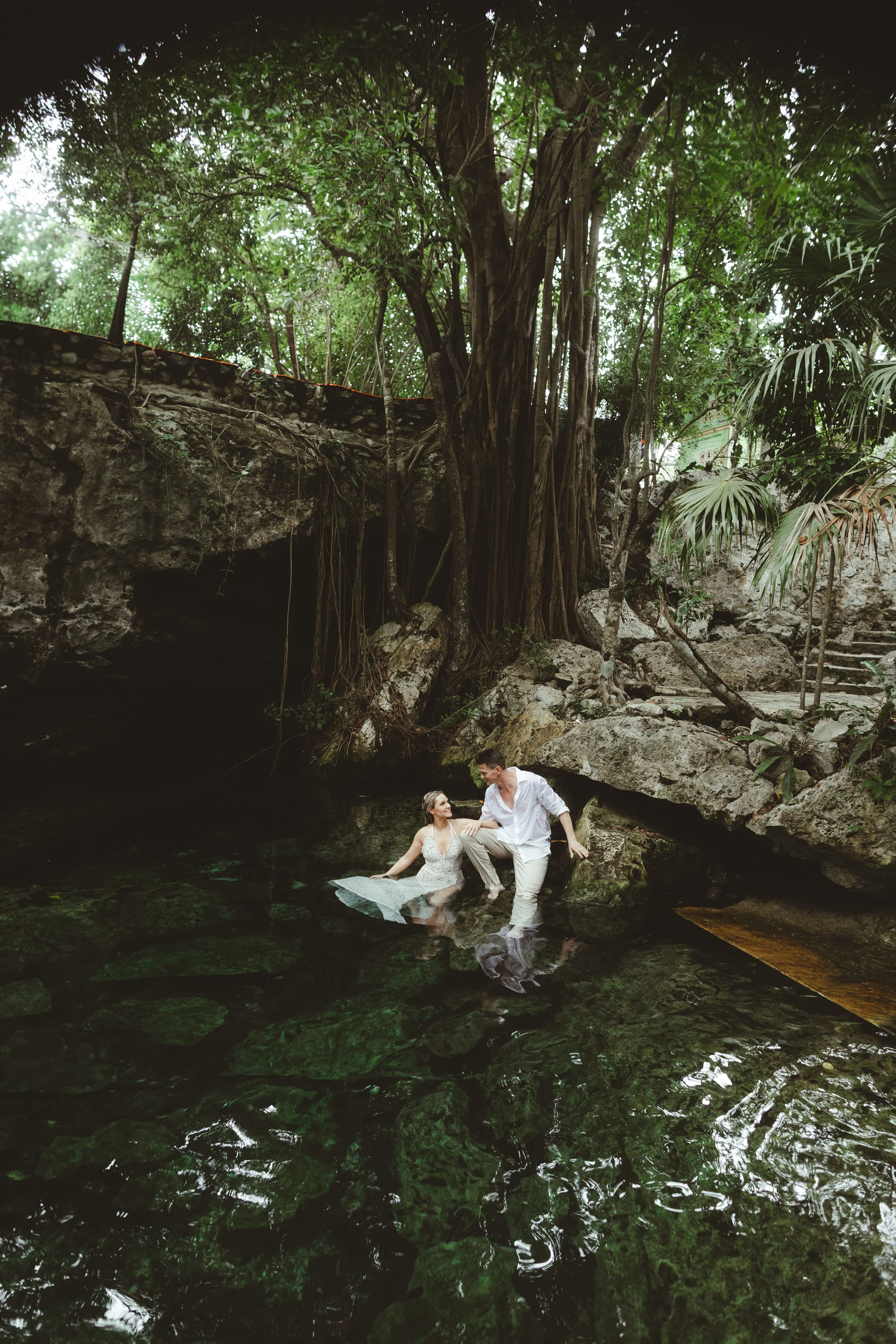 A young couple in wedding attire standing in a shallow cenote surrounded by lush greenery and large trees. Cenote wedding Photoshoot in Playa del Carmen.