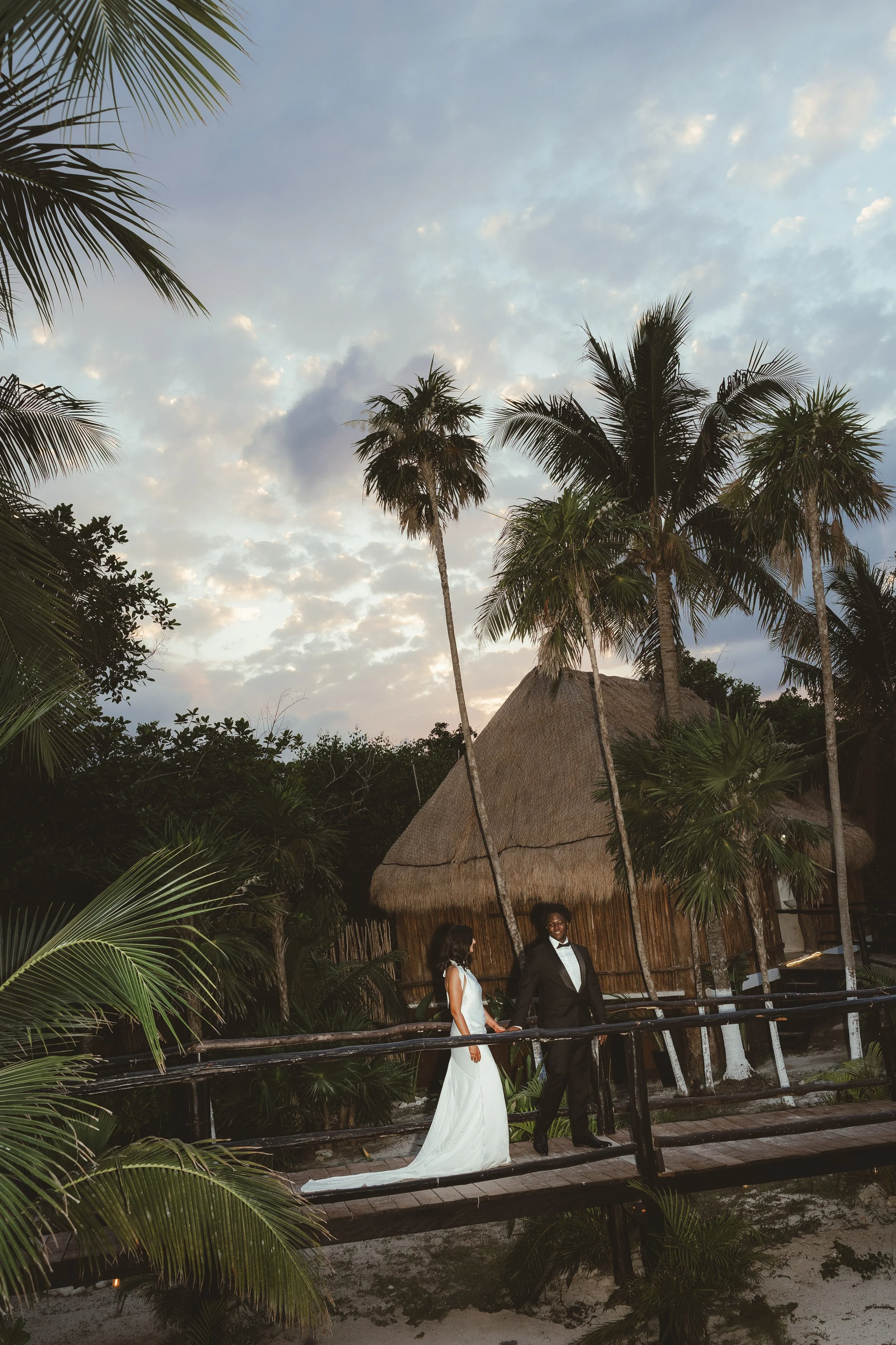 A bride and groom standing on a wooden bridge surrounded by tropical palm trees near a thatched-roof hut during sunset.