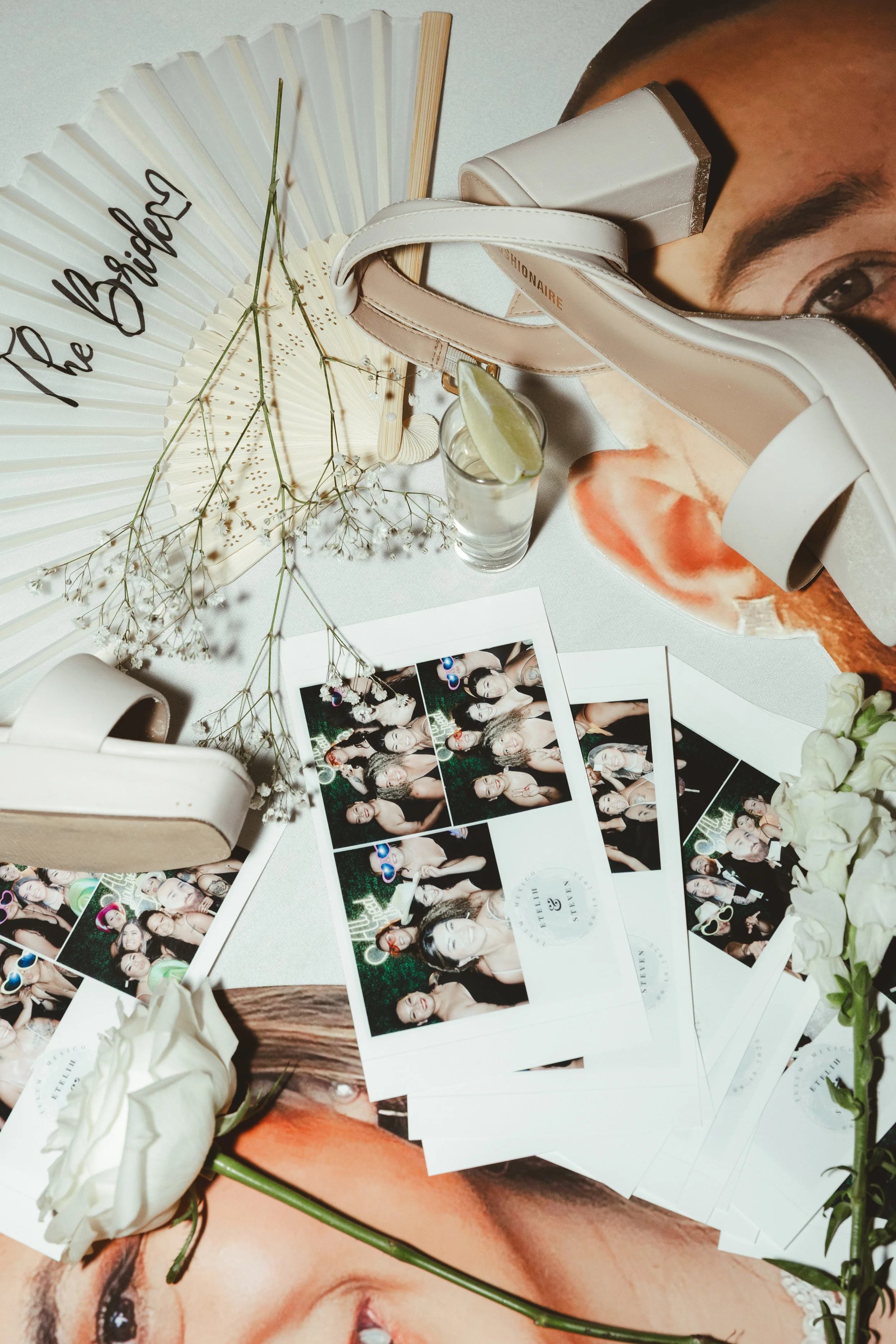 A collection of photo booth pictures with people, a white rose, a paper fan with the words 'the bride,' sunglasses, a glass with a lime wedge, and decorative flowers on a table.
