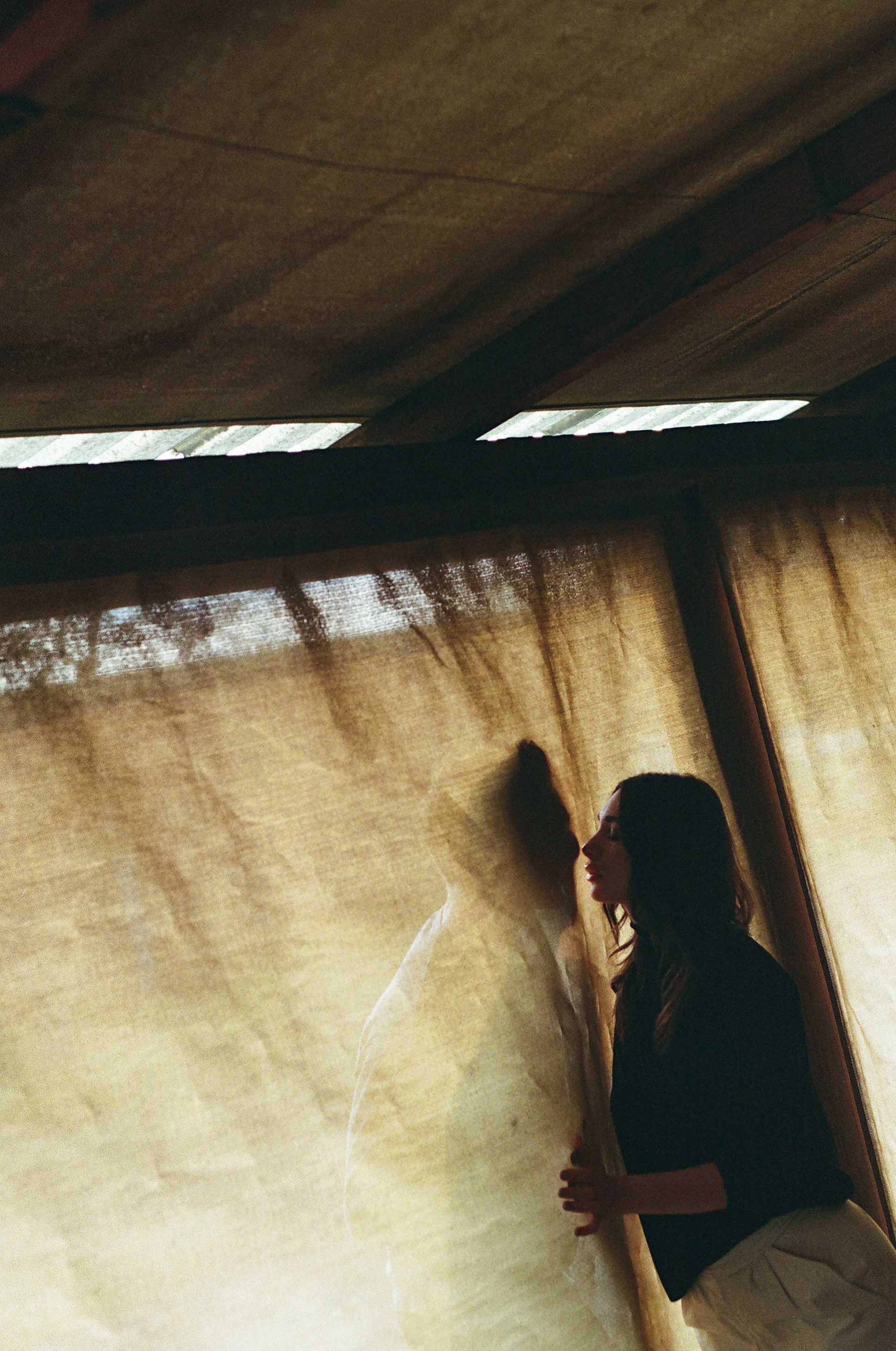 A woman with curly hair and a dark top stands near a textured golden curtain, creating a silhouette with her face in profile against her shadow projected on the curtain.