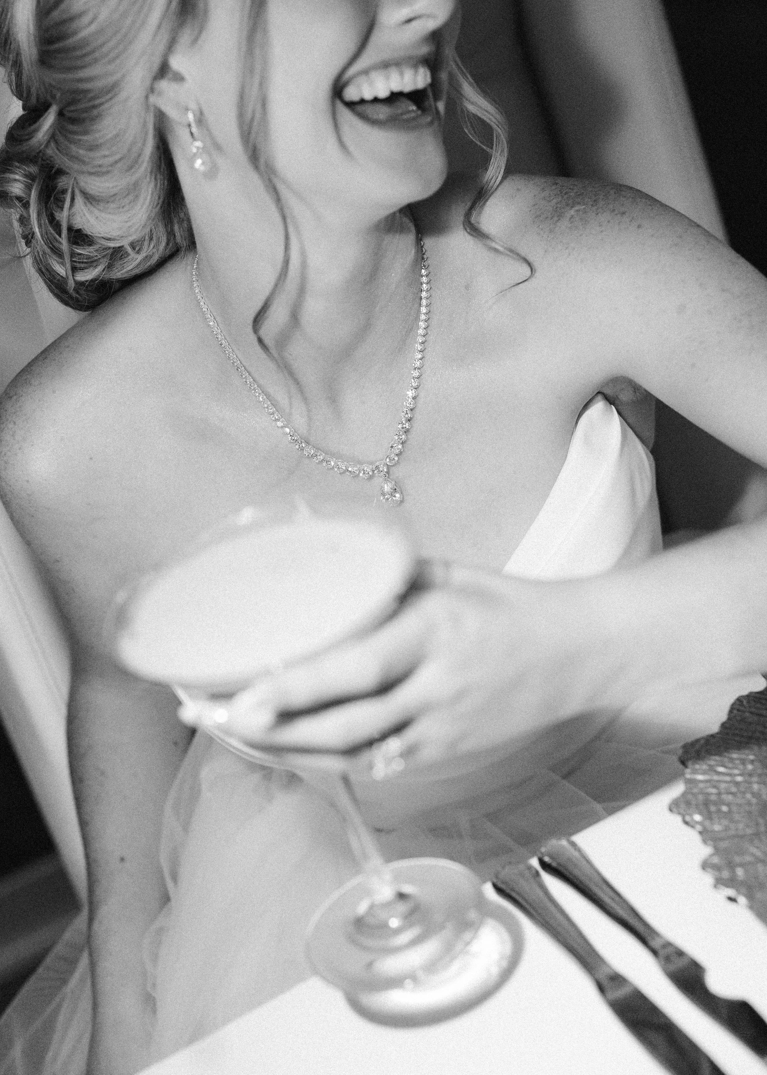 A bride's close up, smiling and holding a glass of expresso martini, with a blurred background. In black and white wedding photograph.