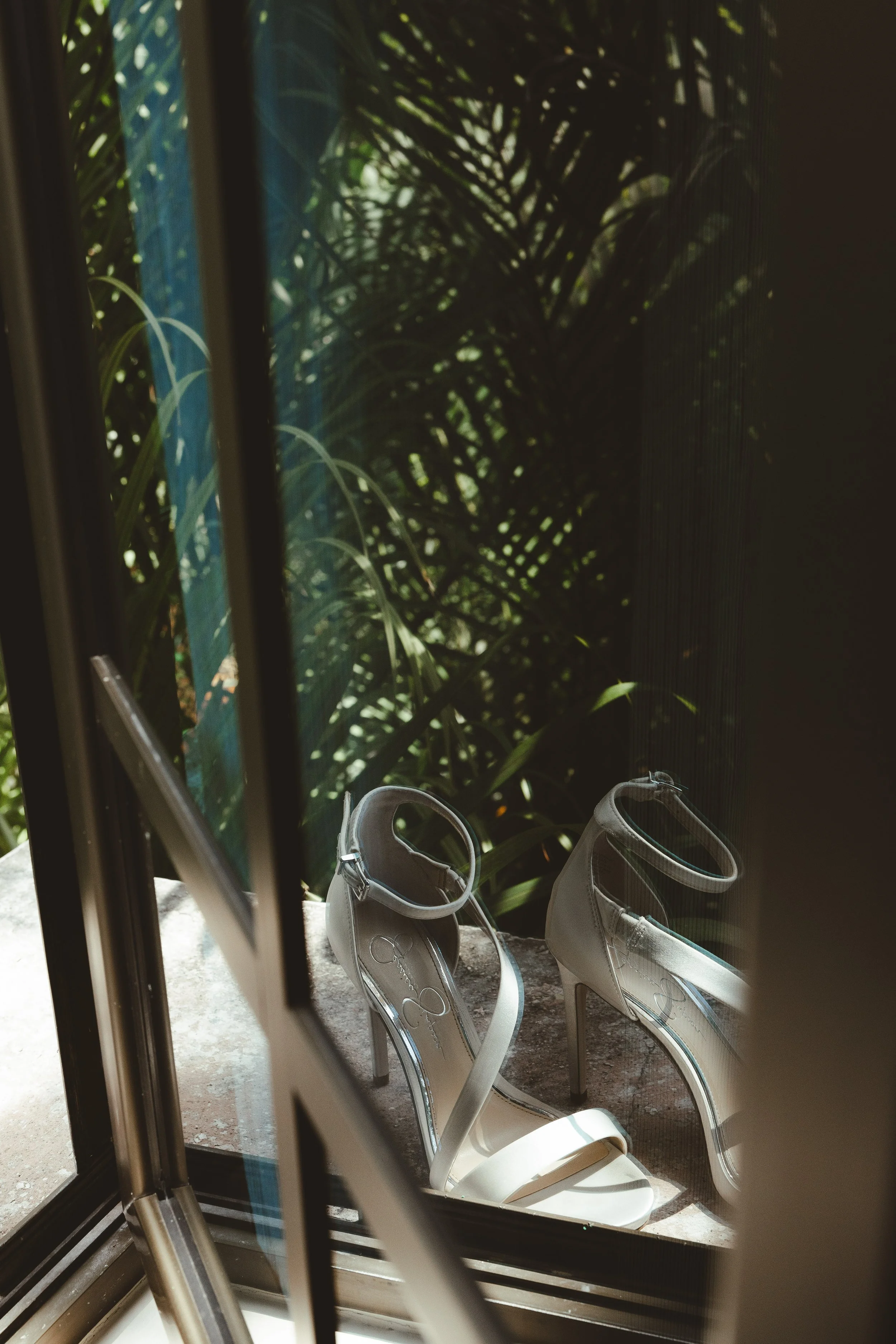 A pair of white high-heeled sandals with ankle straps outside a window, with green plants and sunlight in the background.