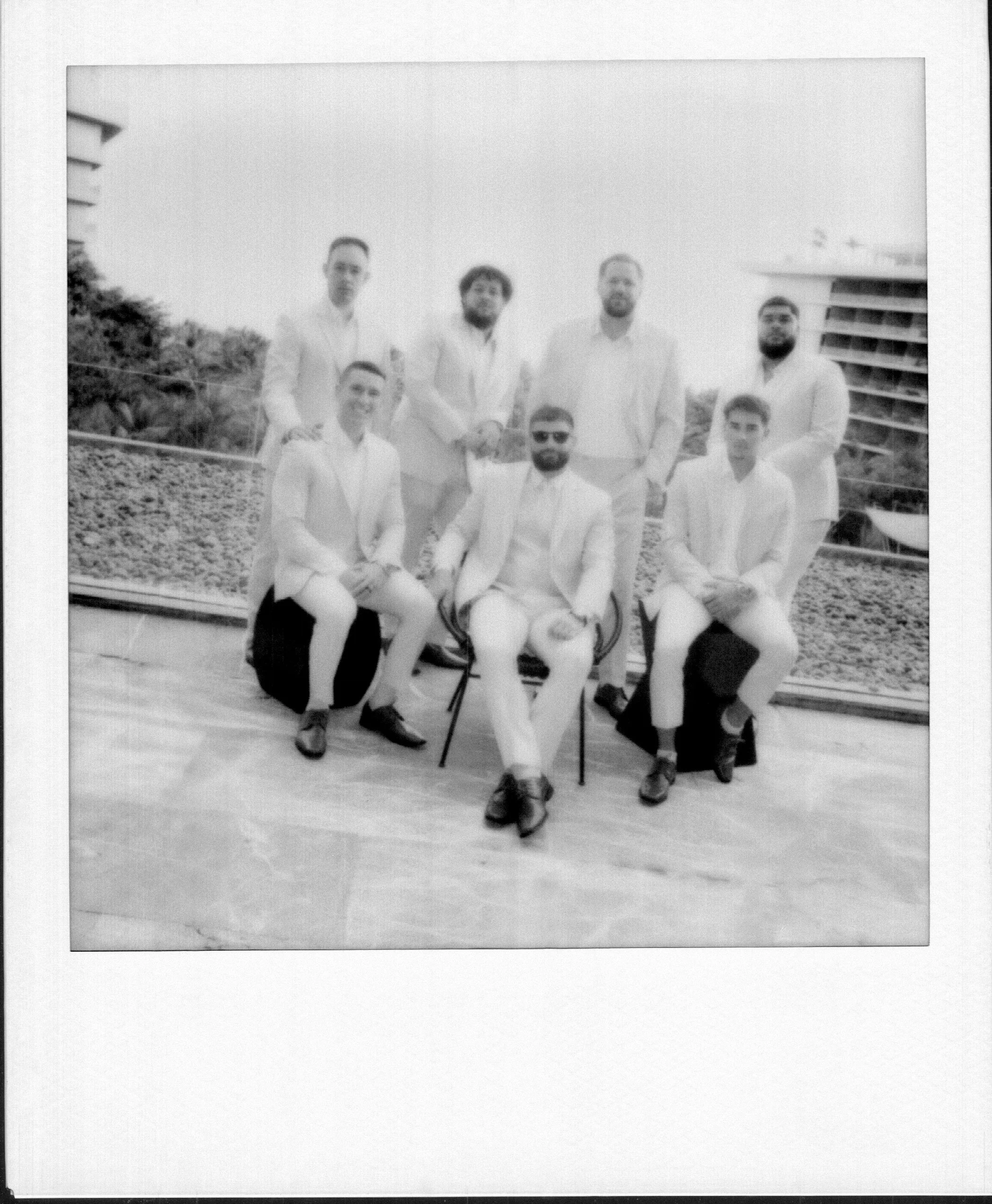 Black and white photo of nine men in white suits, some wearing sunglasses, on a terrace outdoors with a building and trees in the background.