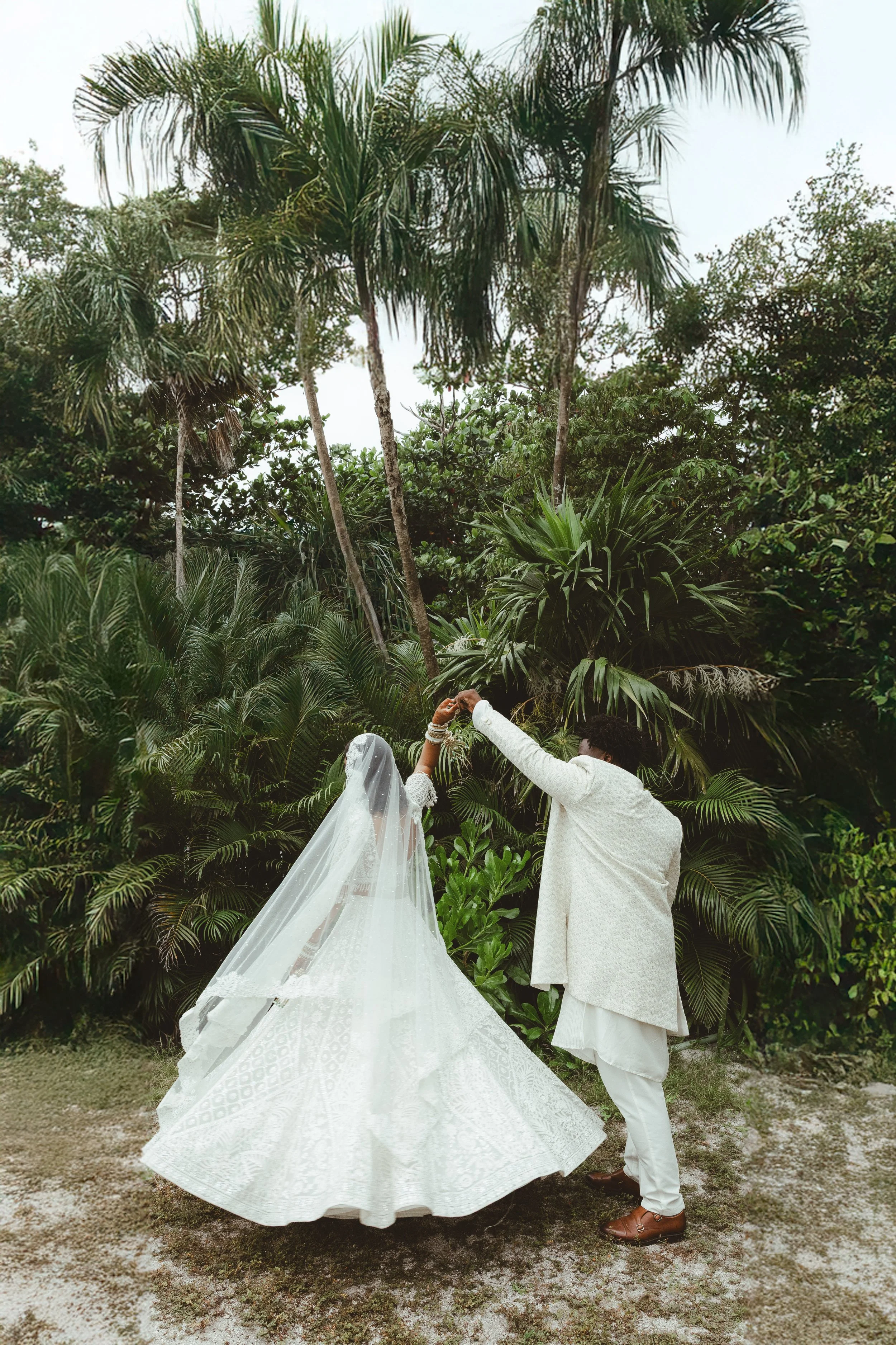 A couple in wedding attire dancing outdoors in front of tropical plants and trees, with the woman wearing a white wedding gown and veil, and the man in a cream-colored traditional outfit.