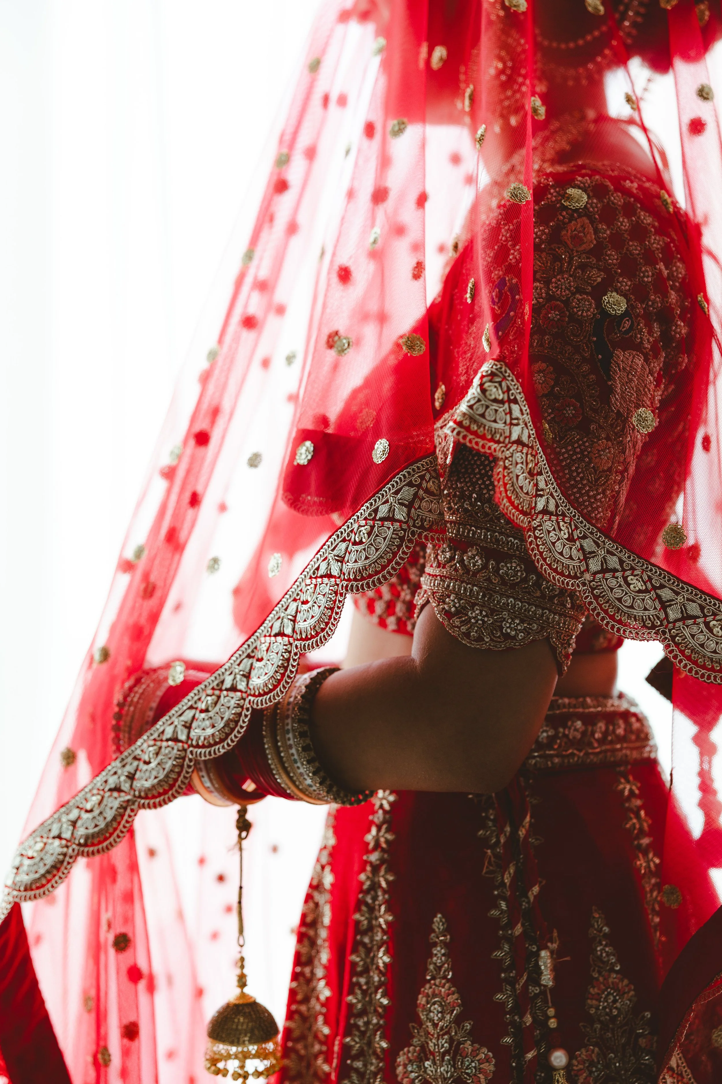 Close-up of a woman wearing a red embroidered traditional Indian outfit, partially covered by a sheer red dupatta with gold embellishments, showing intricate jewelry and henna on her arm.