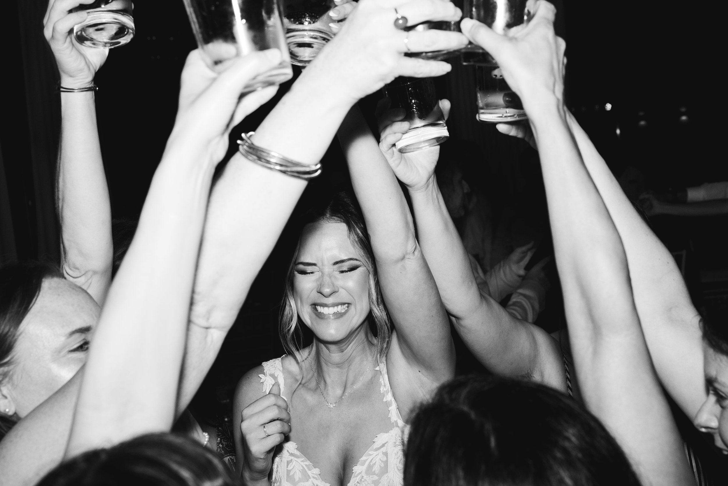 A group of women raising glasses in a toast at a celebration, with one woman smiling and appearing joyful, in a black and white photo during a wedding party in cancun.