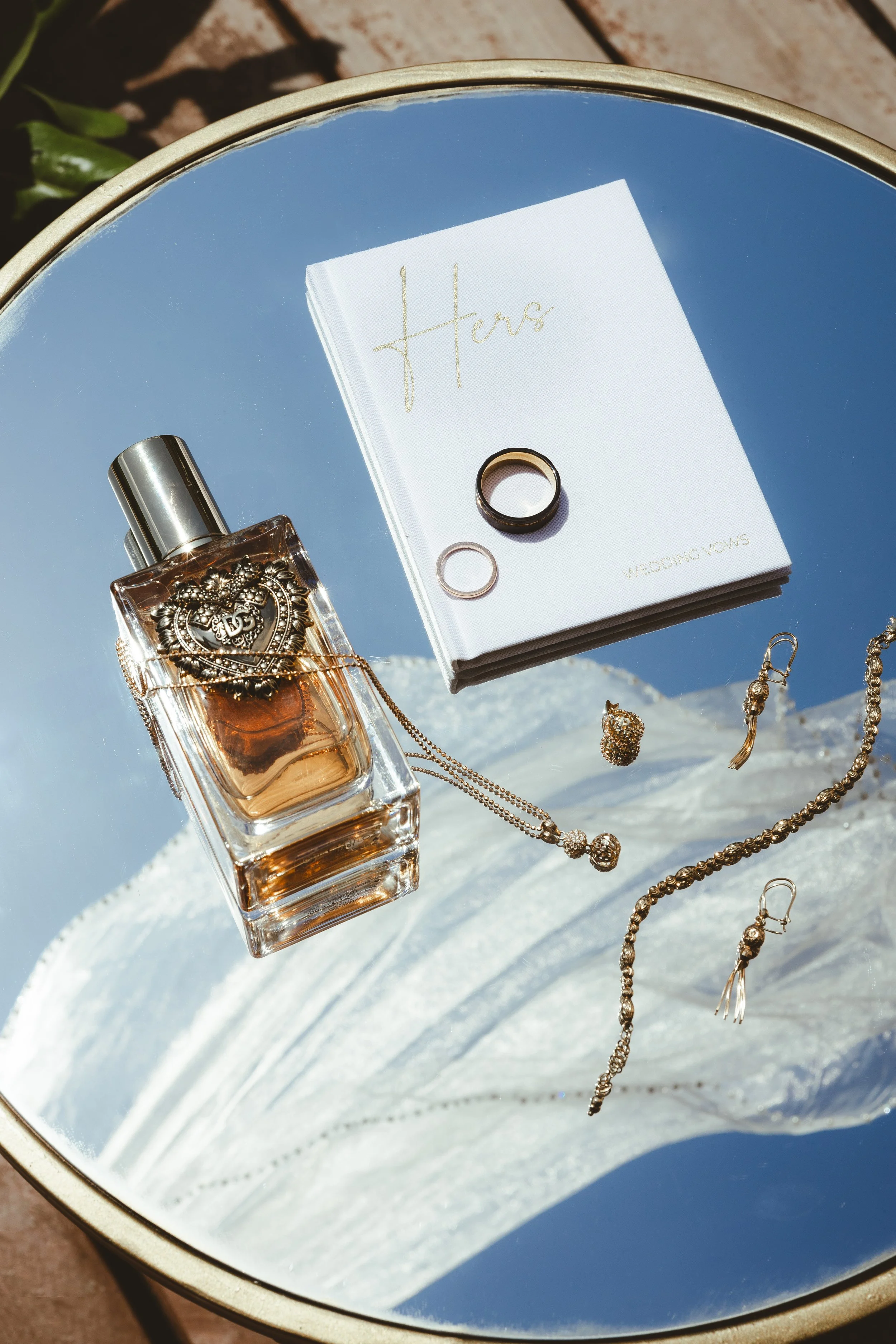 A round side table holding a perfume bottle, jewelry, and a wedding vow booklet labeled "Hers". The jewelry includes rings, earrings, and layered necklaces with decorative beads. The scene is illuminated by natural sunlight.