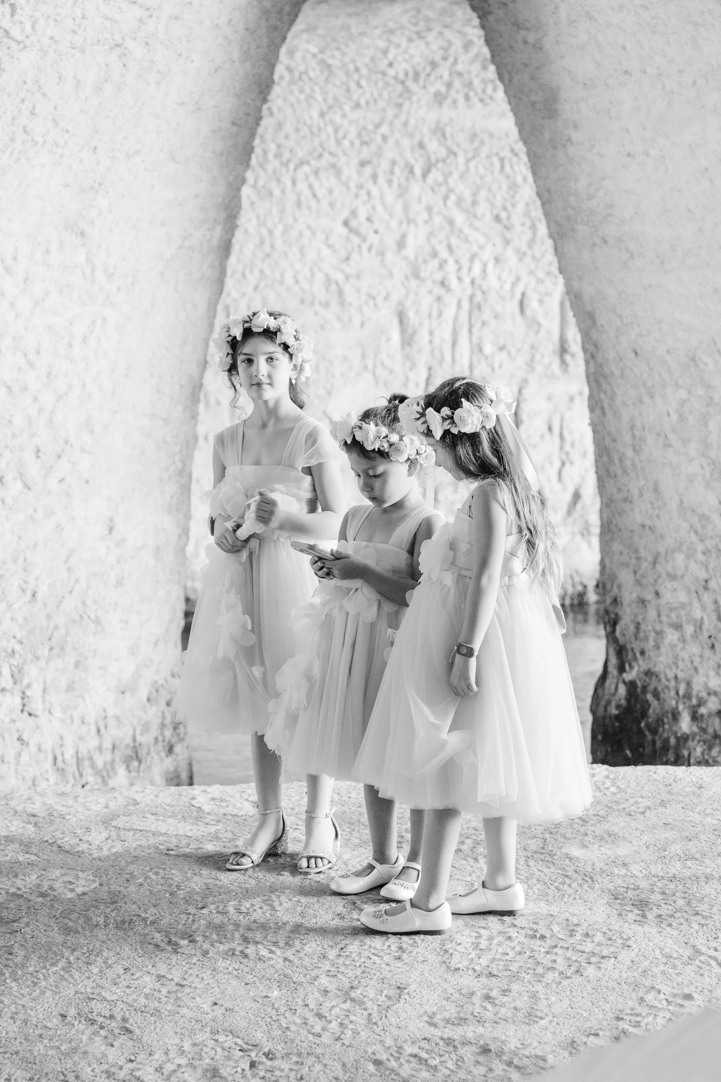 Black and white photo of three young girls dressed in flower girl dresses with floral crowns, standing on a gravel path against a wall with large trees in the background in Xcaret Mexico Resort Wedding.