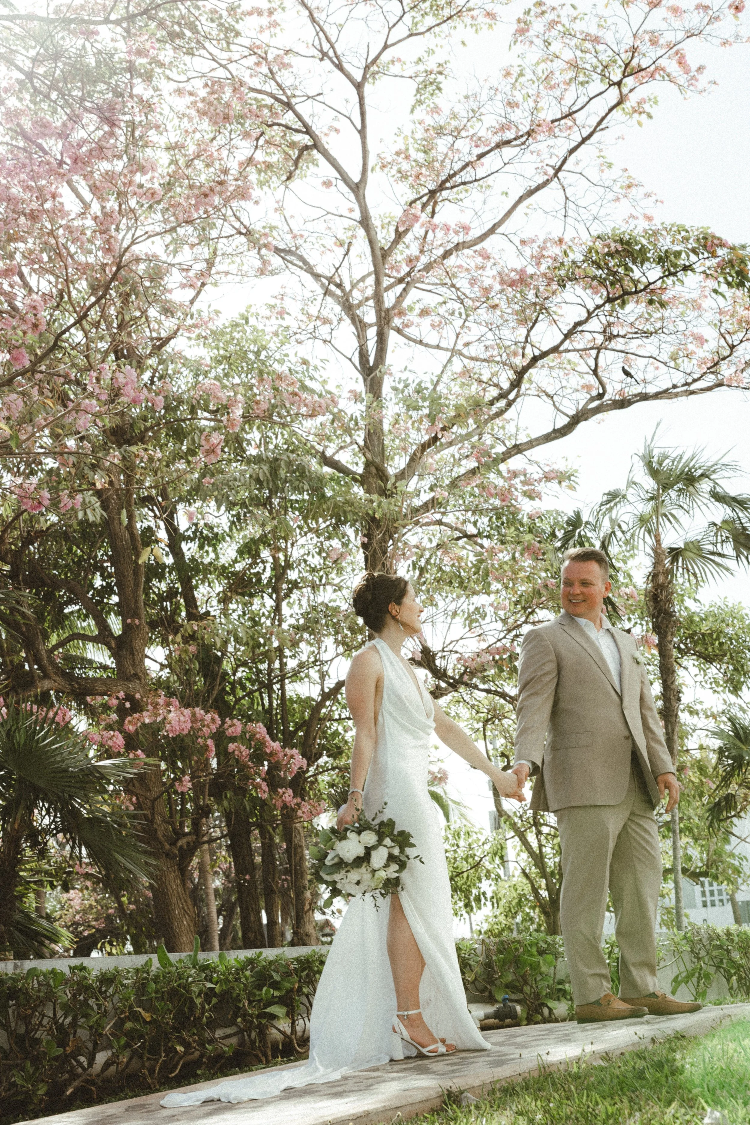 A bride and groom holding hands during their wedding ceremony outdoors among pink flowering trees, with lush greenery and palm plants in the background in Cancun.