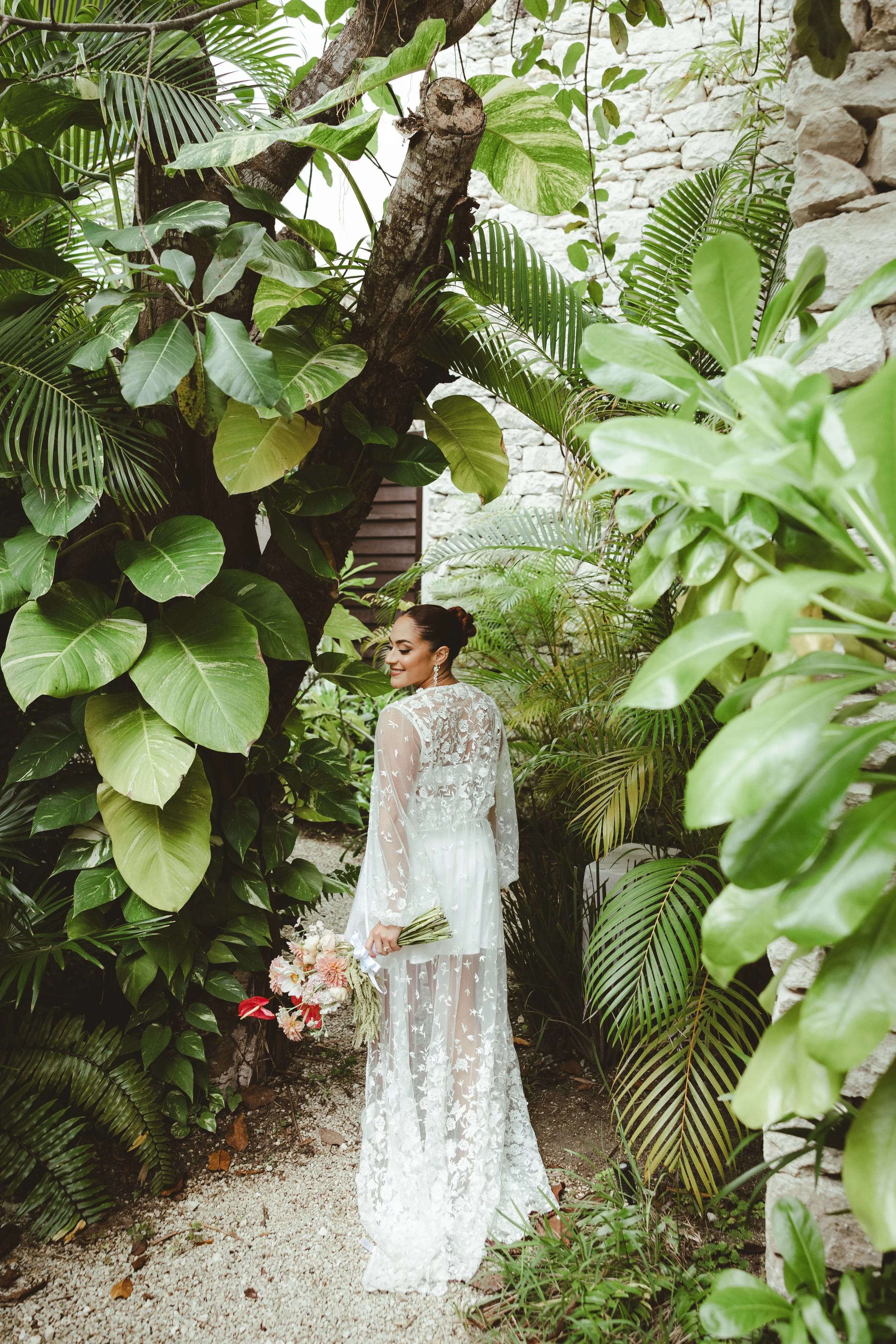 Bride in a white wedding dress surrounded by tropical plants and greenery, smiling and looking back  in Secrets Moxche Playa del Carmen.