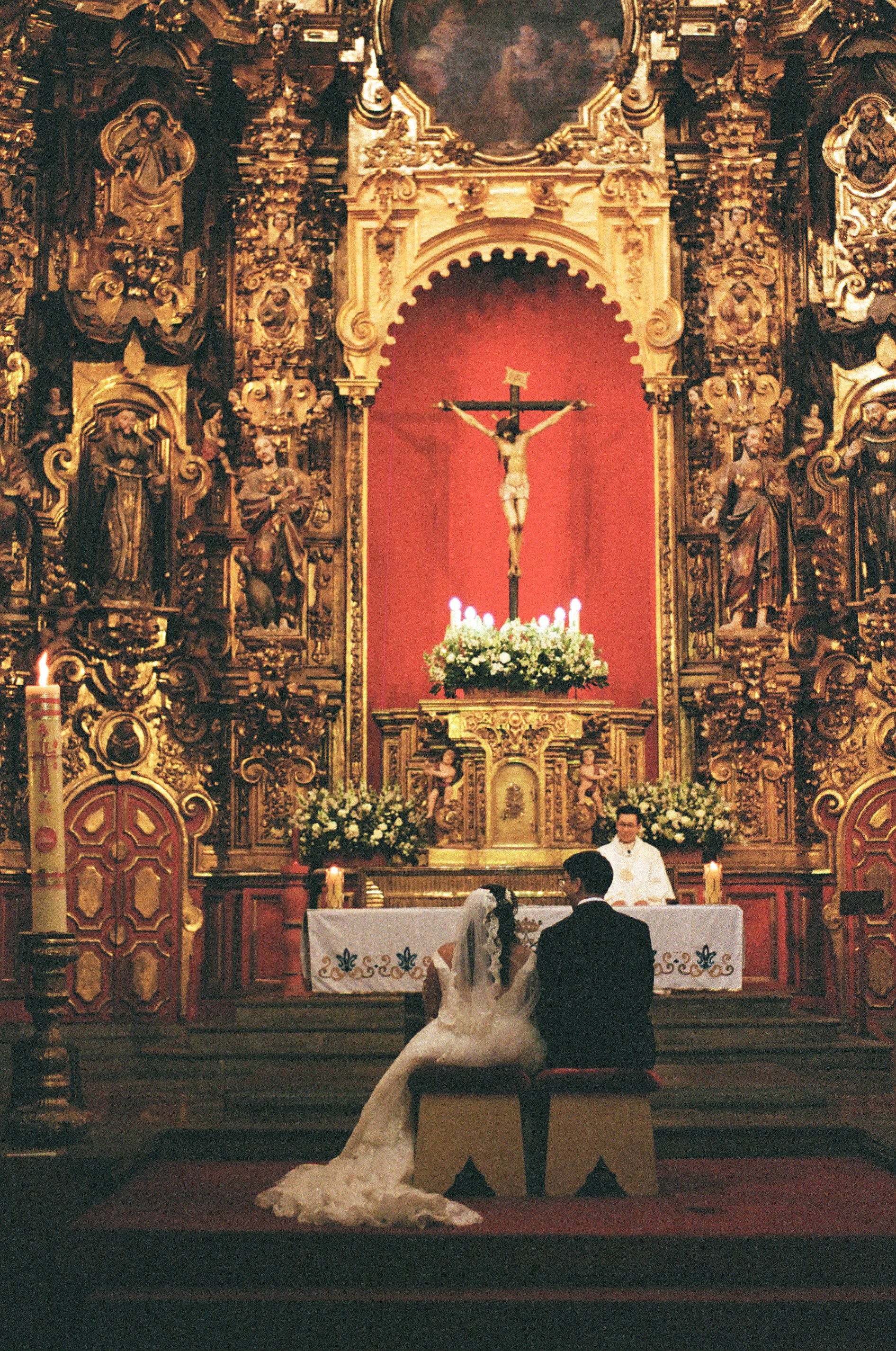 A couple getting married at a church altar with a priest officiating. The bride wears a white wedding dress and veil, and the groom wears a black suit. The altar has ornate gold decorations, religious statues, and a crucifix with Jesus on the cross w